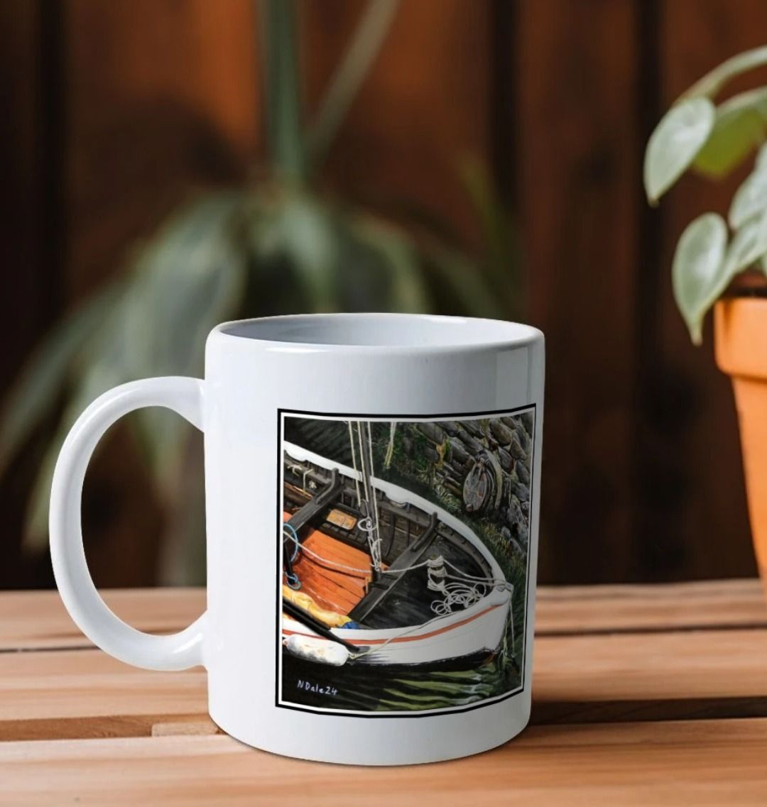 Image of a White Mug on a wooden shelf featuring a copy of the painting 'The Outlet' by Nick Dale. The handle is orientated to the left. The mug depicts a view down into the harbour at Lynmouth in North Devon showing a small sailing boat at rest against the harbour wall next to a water outfall pipe.