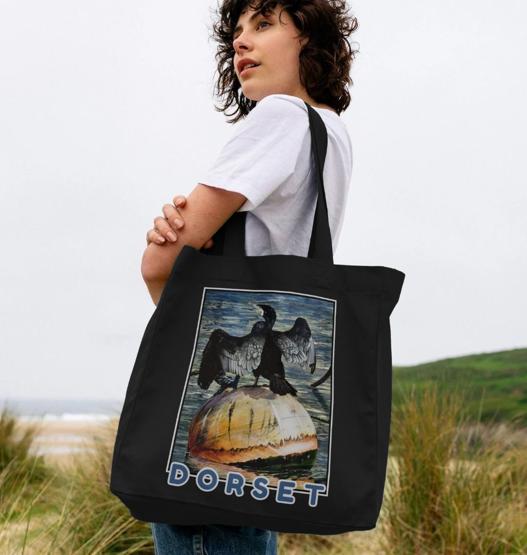 Image of a female model holding a Black Shopper Tote Bag featuring a copy of the painting 'Cormorant At West Bay' by Nick Dale. The bag depicts a close-up of a cormorant sunbathing on a mooring buoy in West Bay's harbour in Dorset.