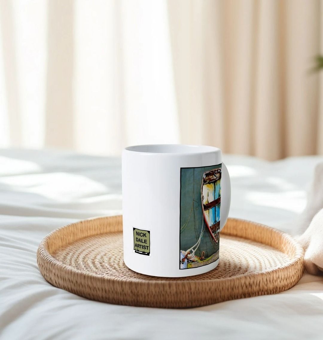 Image of a White Mug on a tray on a bed featuring a copy of the painting 'Good Intentions at Lyme Regis' by Nick Dale. Image also shows artist's logo on the mug. The mug handle is obscured behind the mug. The mug depicts a small wooden rowing boat tied up in Lyme Regis harbour, Dorset.