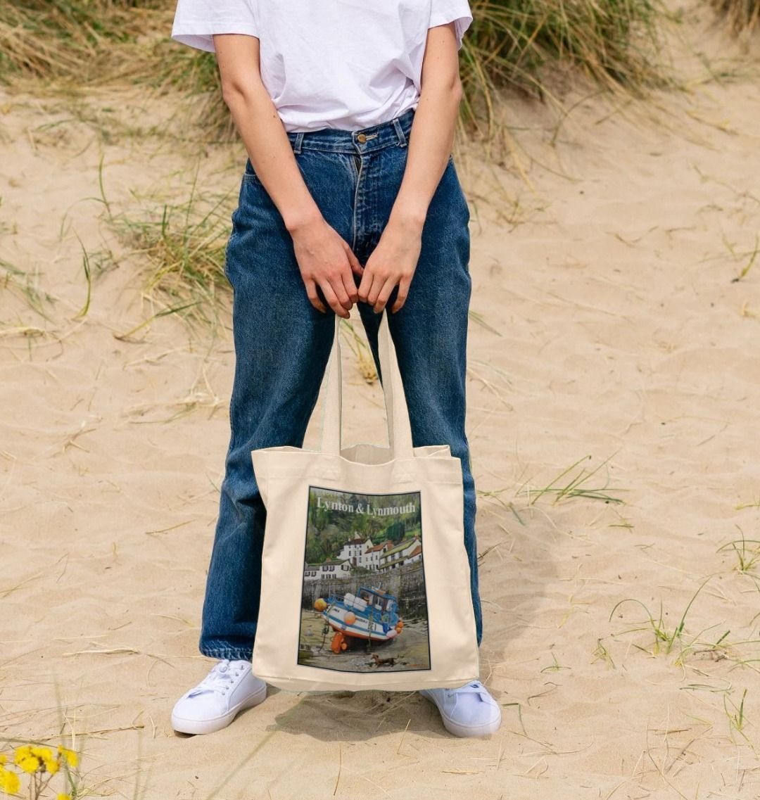 Image of a female model holding a Natural Shopper Tote Bag featuring a copy of the painting 'Lynmouth At Lowest Tide' by Nick Dale, with the words 'Lynton & Lynmouth' written across the top. The bag depicts an unusual perspective on the picturesque harbour of Lynmouth (Devon), as seen from the floor of the harbour at low tide. Behind the fishing boat can be seen the characterful harbourside buildings.