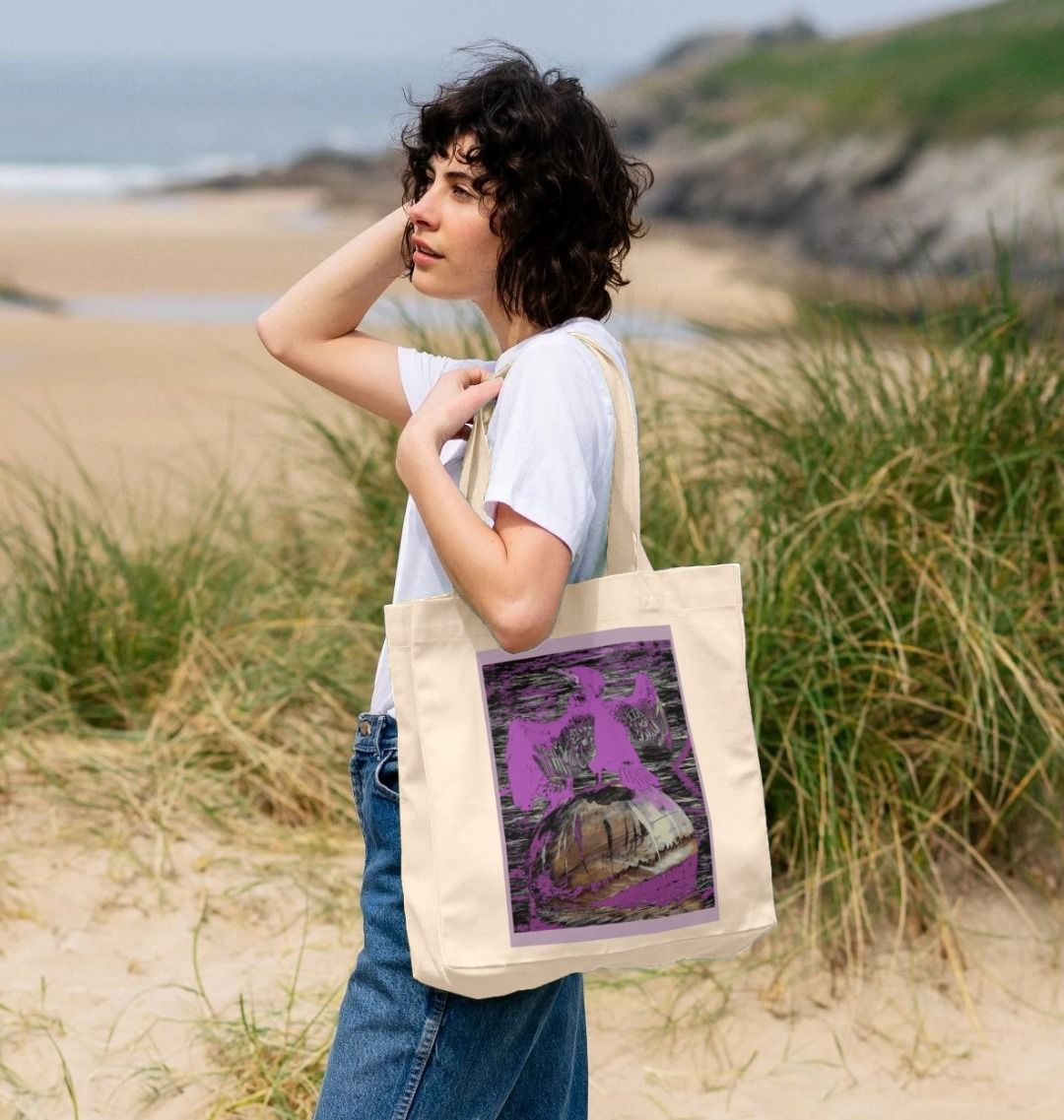 Image of a female model holding a Natural Shopper Tote Bag featuring a modified copy of the painting 'Cormorant At West Bay' by Nick Dale. The bag depicts a close-up of a cormorant sunbathing on a mooring buoy in West Bay's harbour in Dorset.