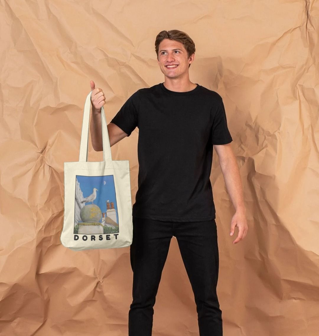 Image of male model holding Natural Tote Bag featuring a copy of the painting 'A View Of The Sea At Lyme Regis' by Nick Dale. The bag depicts a gull and buildings along Marine Parade, Lyme Regis.