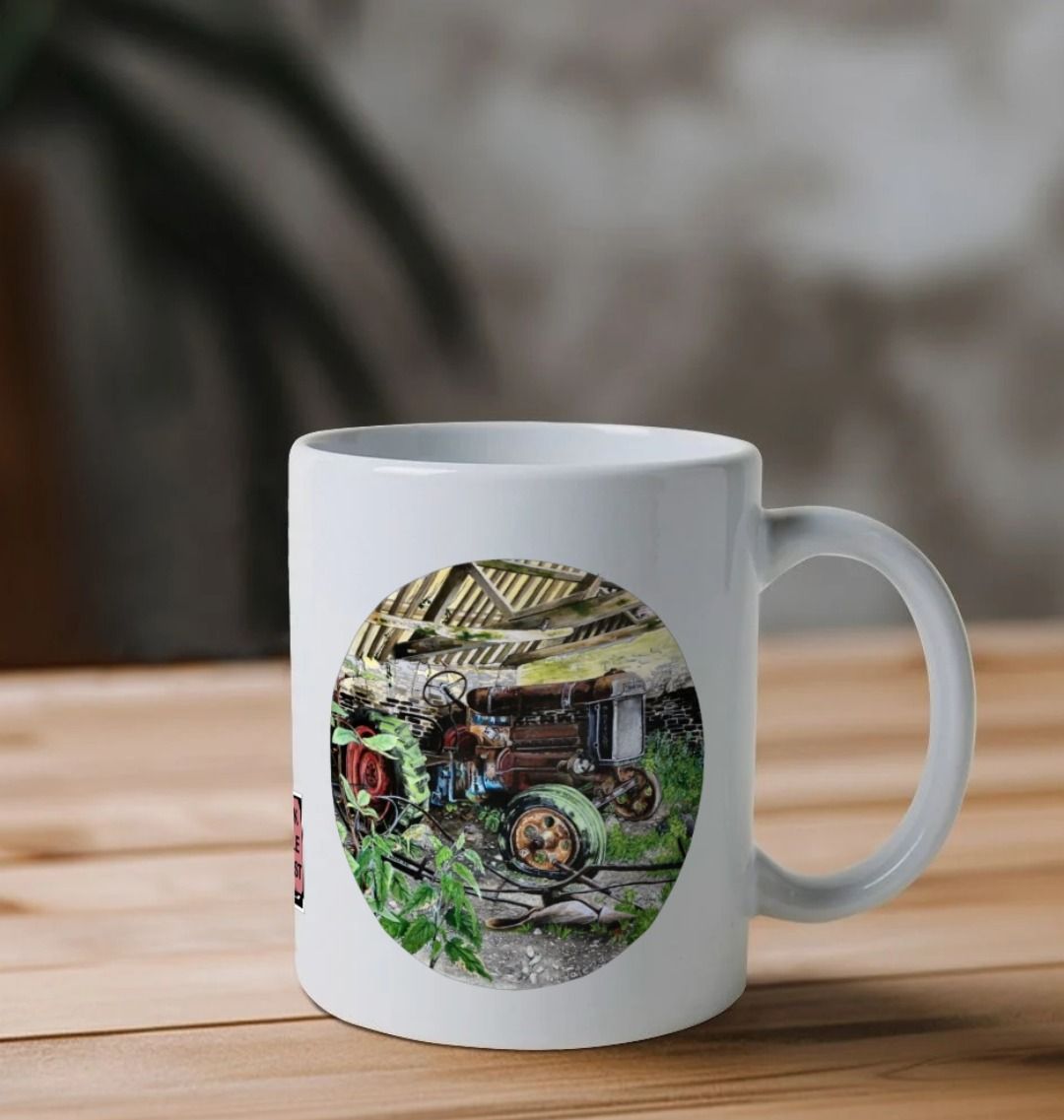 Image of a White Mug on a table featuring a copy of the painting 'A Well Deserved Rest At Tyneham' by Nick Dale. The handle is orientated to the right. The mug depicts a Fordson Major tractor laid up in a farm building at the abandoned village of Tyneham in Dorset.