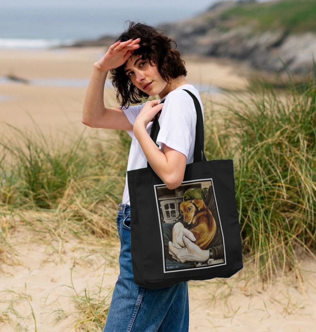Image of a female model holding a Black Shopper Tote Bag featuring a copy of the painting 'Amber Concordia' by Nick Dale. The bag depicts a dog sitting curled-up on the lap of its owner in front of the hearth.