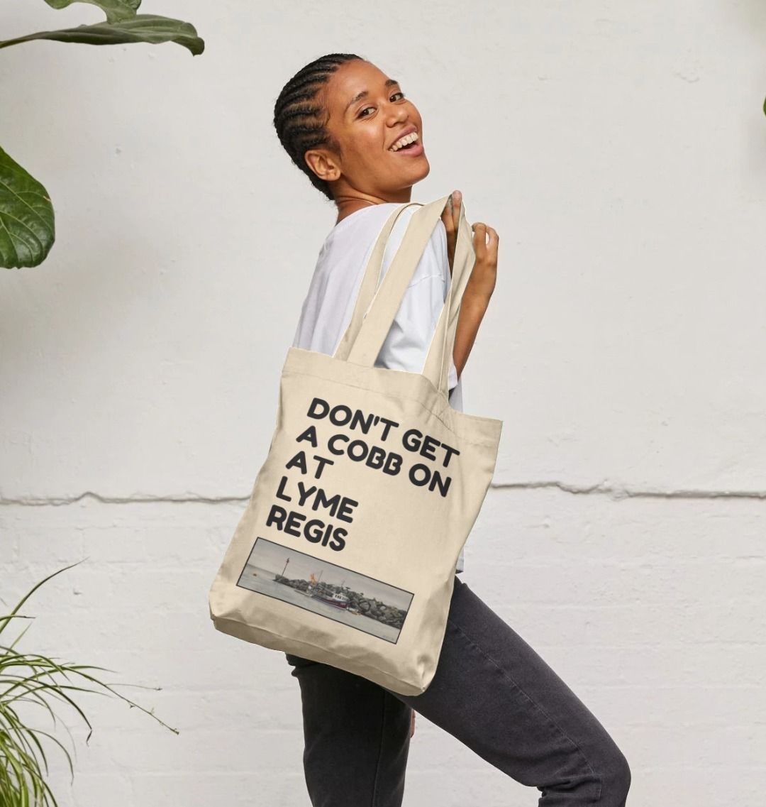 Image of a female model carrying a Natural Tote Bag featuring a copy of the painting 'End Of The Line' by Nick Dale with the slogan 'Don't Get A Cobb On At Lyme Regis' written above it. The bag depicts a fisherman preparing his catch for unloading at Lyme Regis as the birds line up on the breakwater and patiently await their turn to feed.