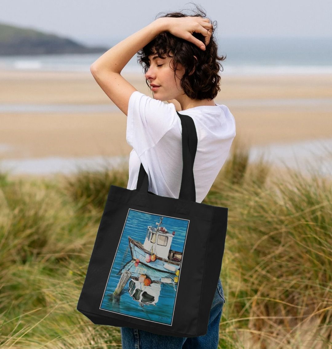 Image of a female model carrying a Black Shopper Tote Bag featuring a copy of the painting 'God's Own 99 Flake Pierces The Veil' by Nick Dale. The bag depicts a fishing boat at rest within the harbour of Lyme Regis, Dorset. The title is based on the similarity of the wooden pile to a chocolate flake as inserted into an ice cream cone. This seemed appropriate given the draw of the town to tourists.