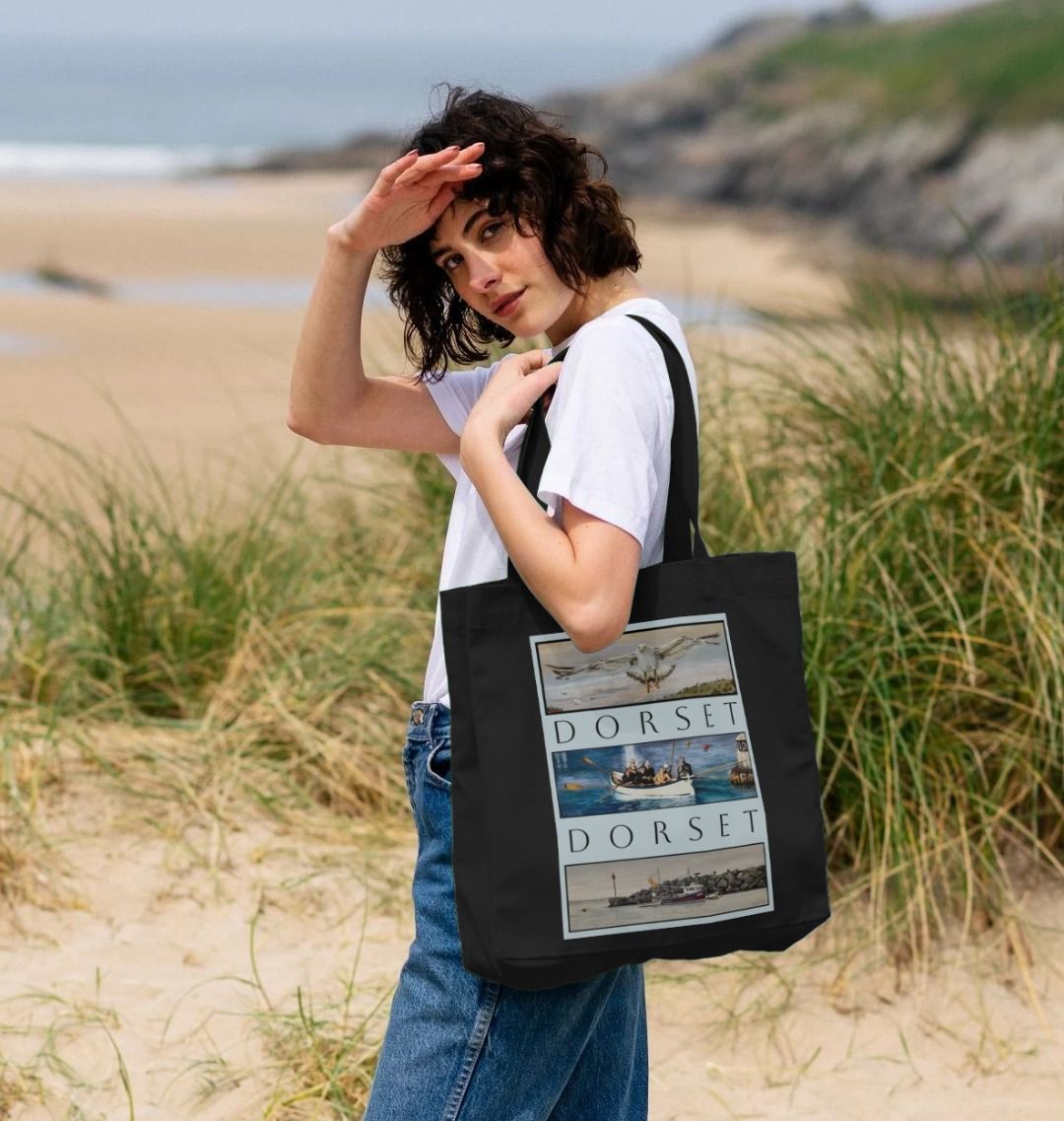 Image of female model holding Black Shopper Tote Bag featuring 3 Scenes Of Dorset. The scenes are derived from 3 original paintings by Nick Dale and are separated from each other vertically by the word 'Dorset'.