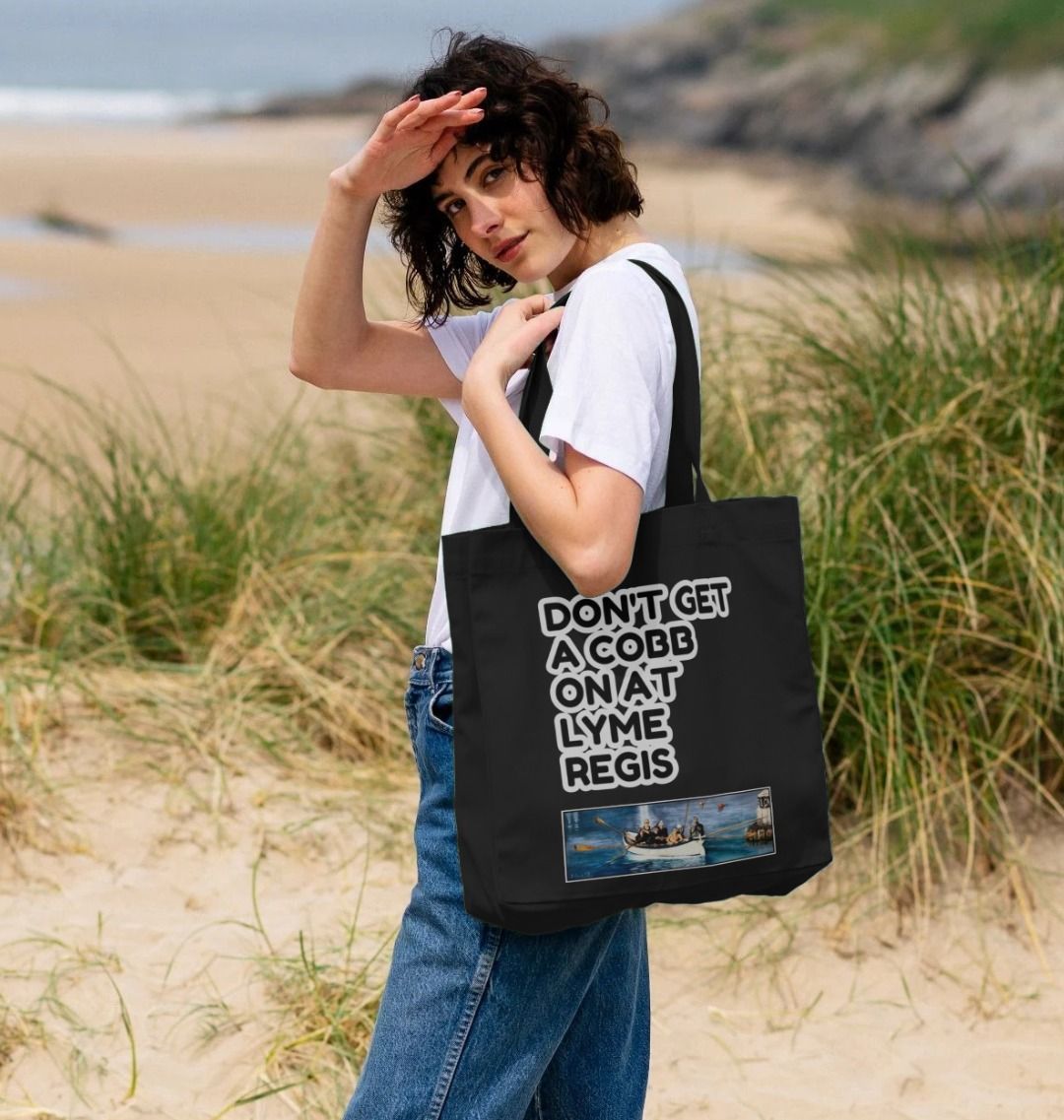 Image of female model carrying a Black Shopper Tote Bag featuring a copy of the painting 'The Return to Base at Lyme Regis' by Nick Dale with the slogan 'Don't Get A Cobb On At Lyme Regis' written above it. The bag depicts the crew of a gig rowing boat returning to Lyme Regis Harbour to take a well-earned rest.