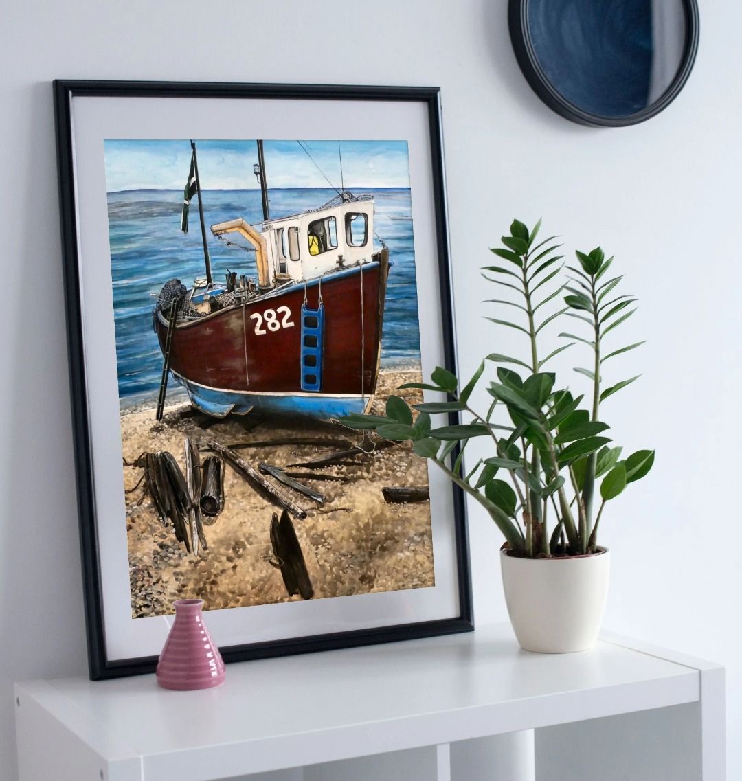 Image of a Large Art Print (A3 size) featuring a copy of the painting 'Relaxing Beer' by Nick Dale. As displayed in a frame on a shelf. The scene depicts a fishing boat beached at Beer, Devon. The wood is laid under the boats to aid in their winching up the shingle beach.