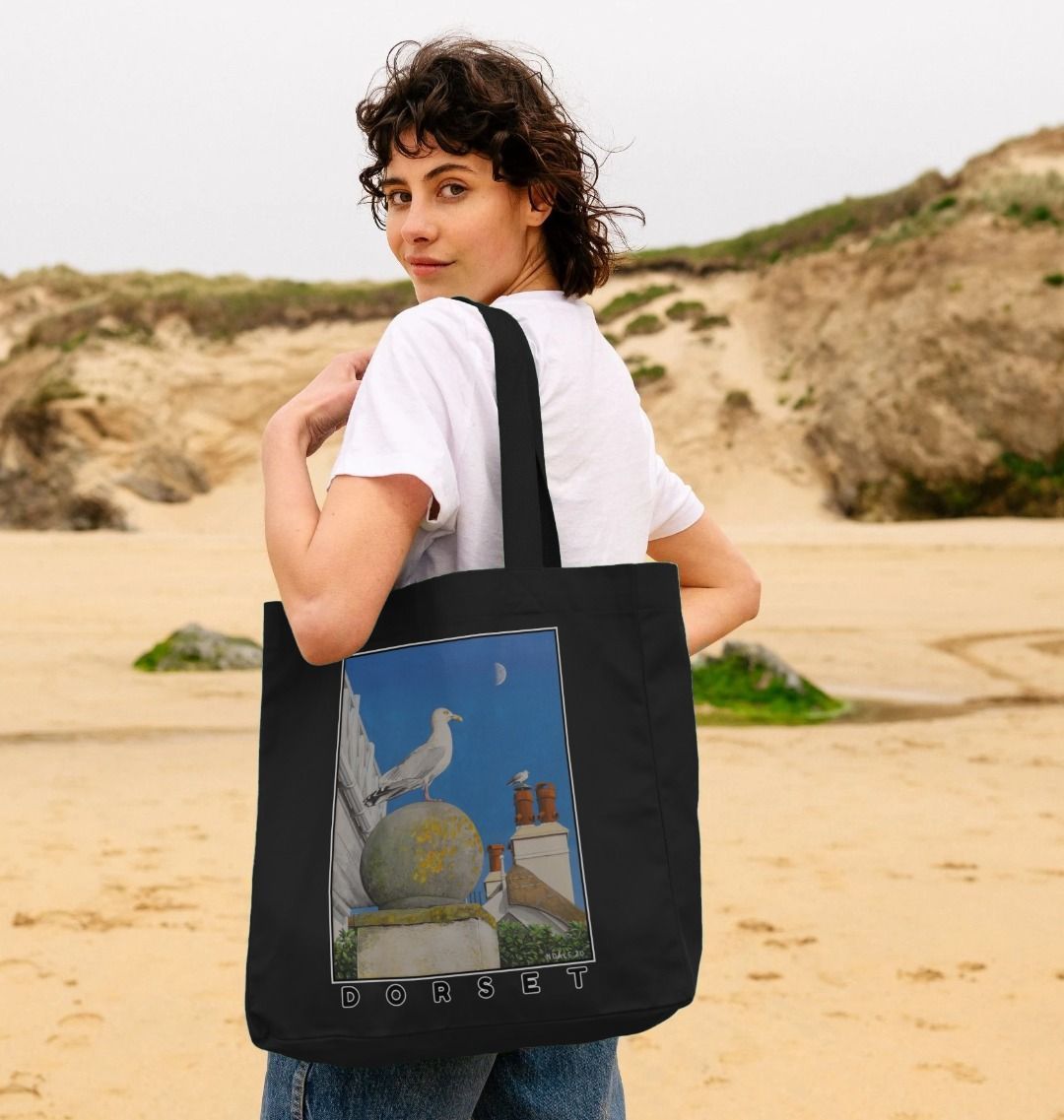Image of female model holding Black Shopper Tote Bag featuring a copy of the painting 'A View Of The Sea At Lyme Regis' by Nick Dale. The bag depicts a gull and buildings along Marine Parade, Lyme Regis.