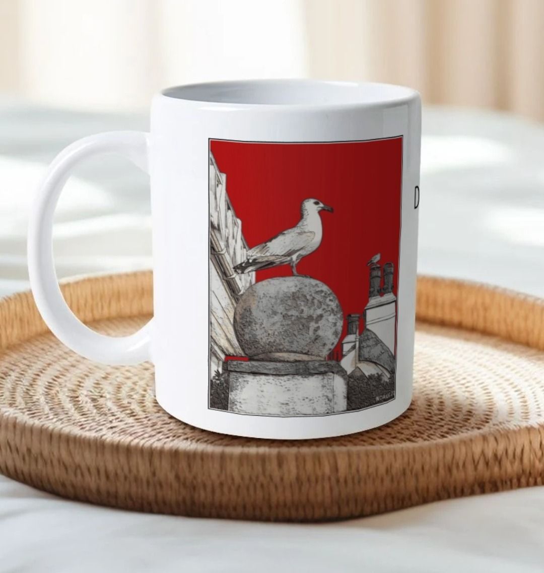 Image of a White Mug on a tray on a bed featuring a modified copy of the painting 'A View Of The Sea At Lyme Regis' by Nick Dale - this being the 'Red' version. The handle is orientated to the left. The mug depicts a gull and buildings along Marine Parade, Lyme Regis.
