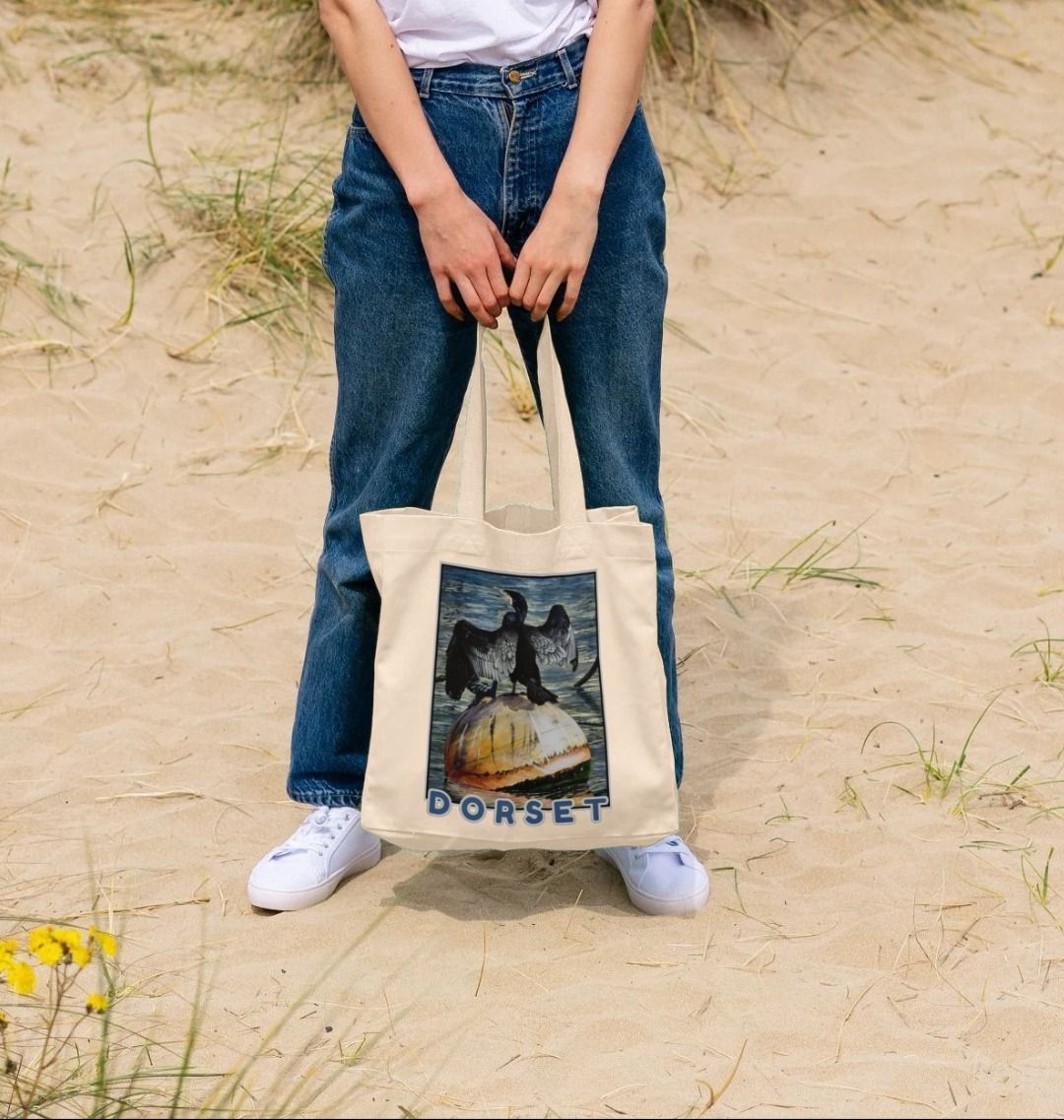 Image of a female model holding a Natural Shopper Tote Bag featuring a copy of the painting 'Cormorant At West Bay' by Nick Dale. The bag depicts a close-up of a cormorant sunbathing on a mooring buoy in West Bay's harbour in Dorset.
