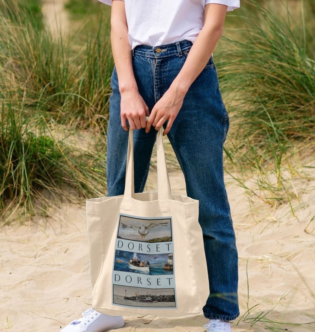 Image of female model holding Natural Shopper Tote Bag featuring 3 Scenes Of Dorset. The scenes are derived from 3 original paintings by Nick Dale and are separated from each other vertically by the word 'Dorset'.