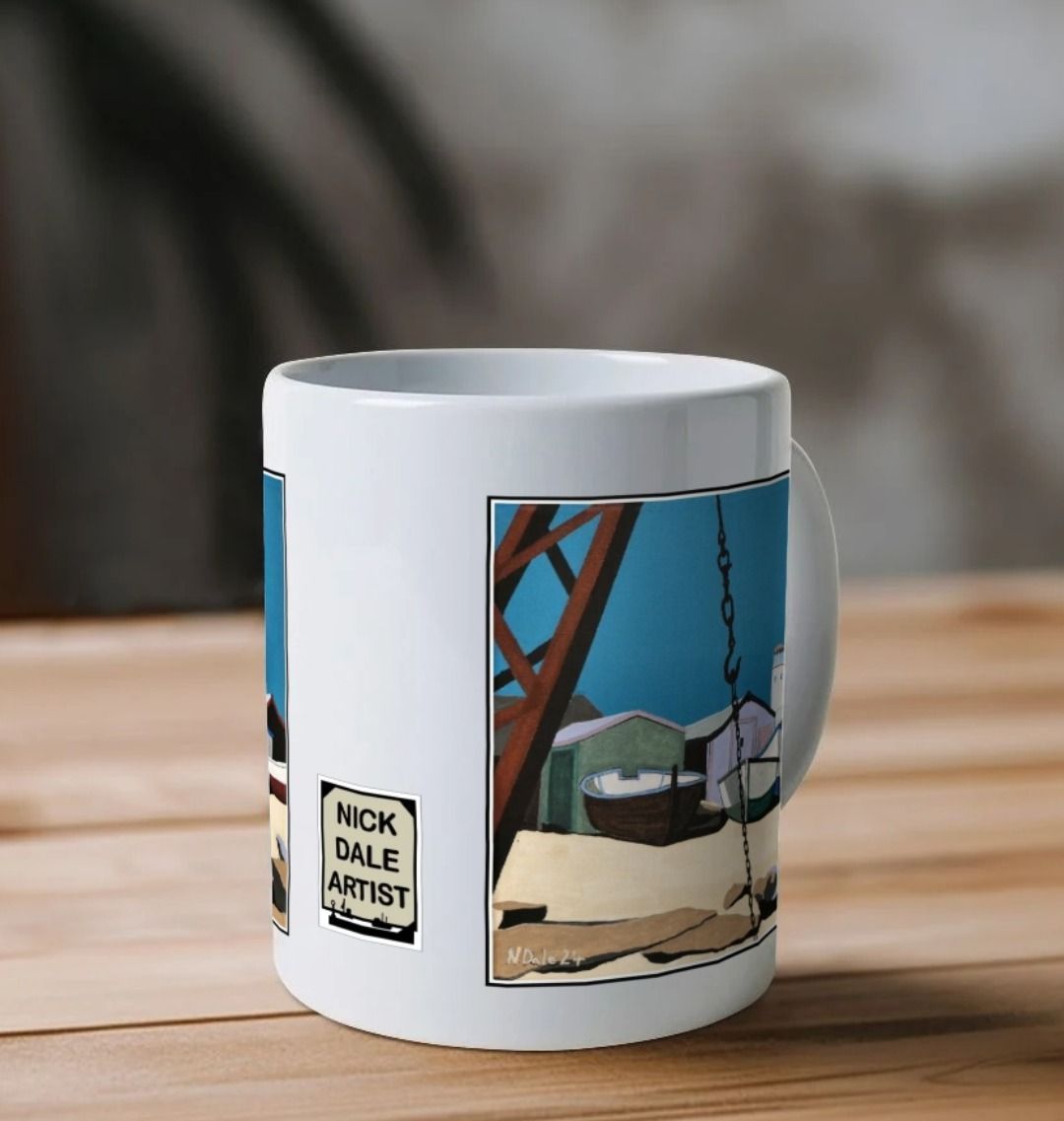 Image of a White Mug on a wooden shelf featuring a copy of the painting 'Old Lower Lighthouse, Portland' by Nick Dale. Image also shows the artist's logo on the mug. The mug handle is obscured behind the mug. The mug depicts the view from the Red Crane at Portland Bill, across to the Old Lower Lighthouse over the beach huts and small boats next to the stone loading quay.