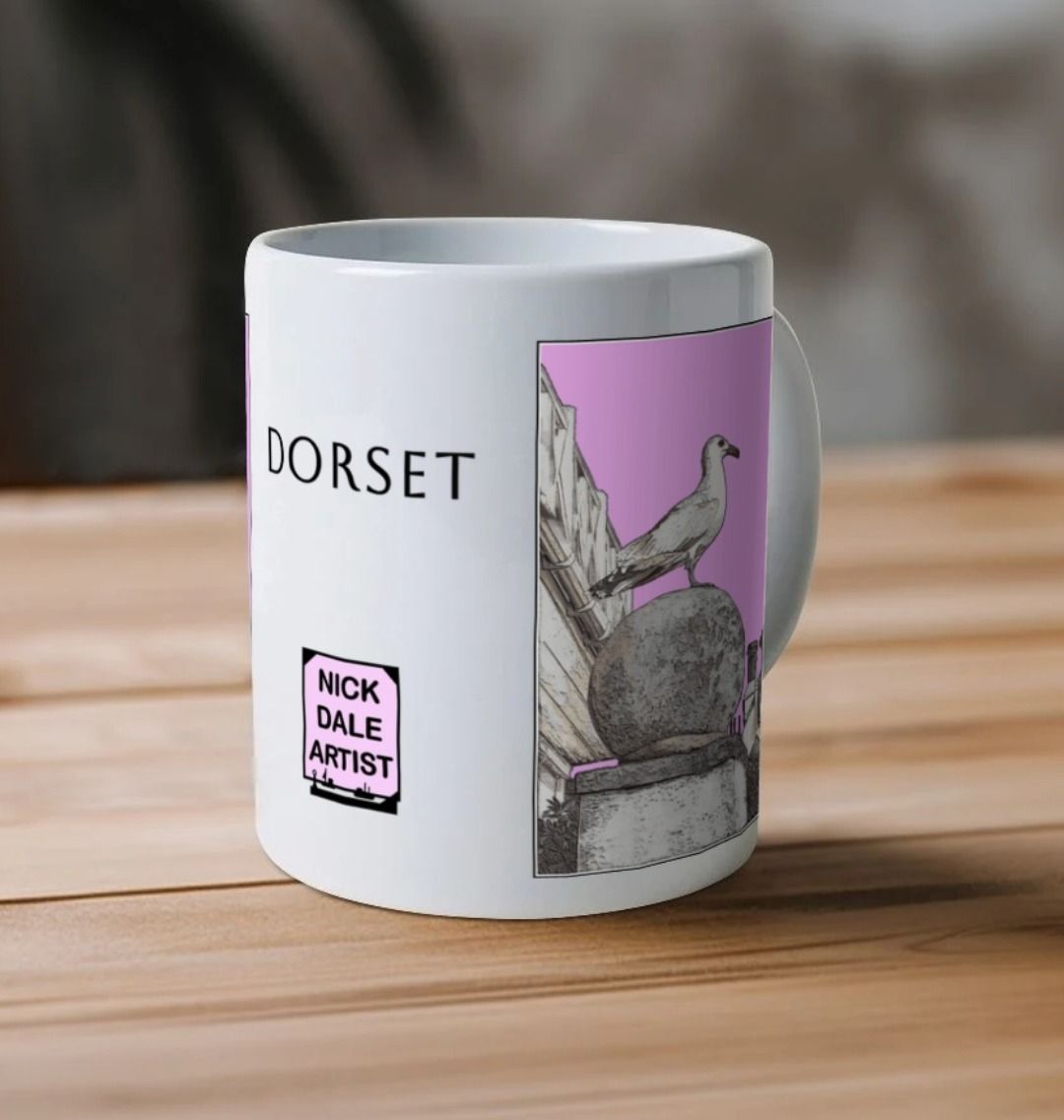 Image of a White Mug on a wooden table featuring a modified copy of the painting 'A View Of The Sea At Lyme Regis' by Nick Dale - this being the 'Pink' version. Image also shows the artist's logo on the mug with the word 'Dorset' written above it. The mug handle is obscured behind the mug. The mug depicts a gull and buildings along Marine Parade, Lyme Regis.