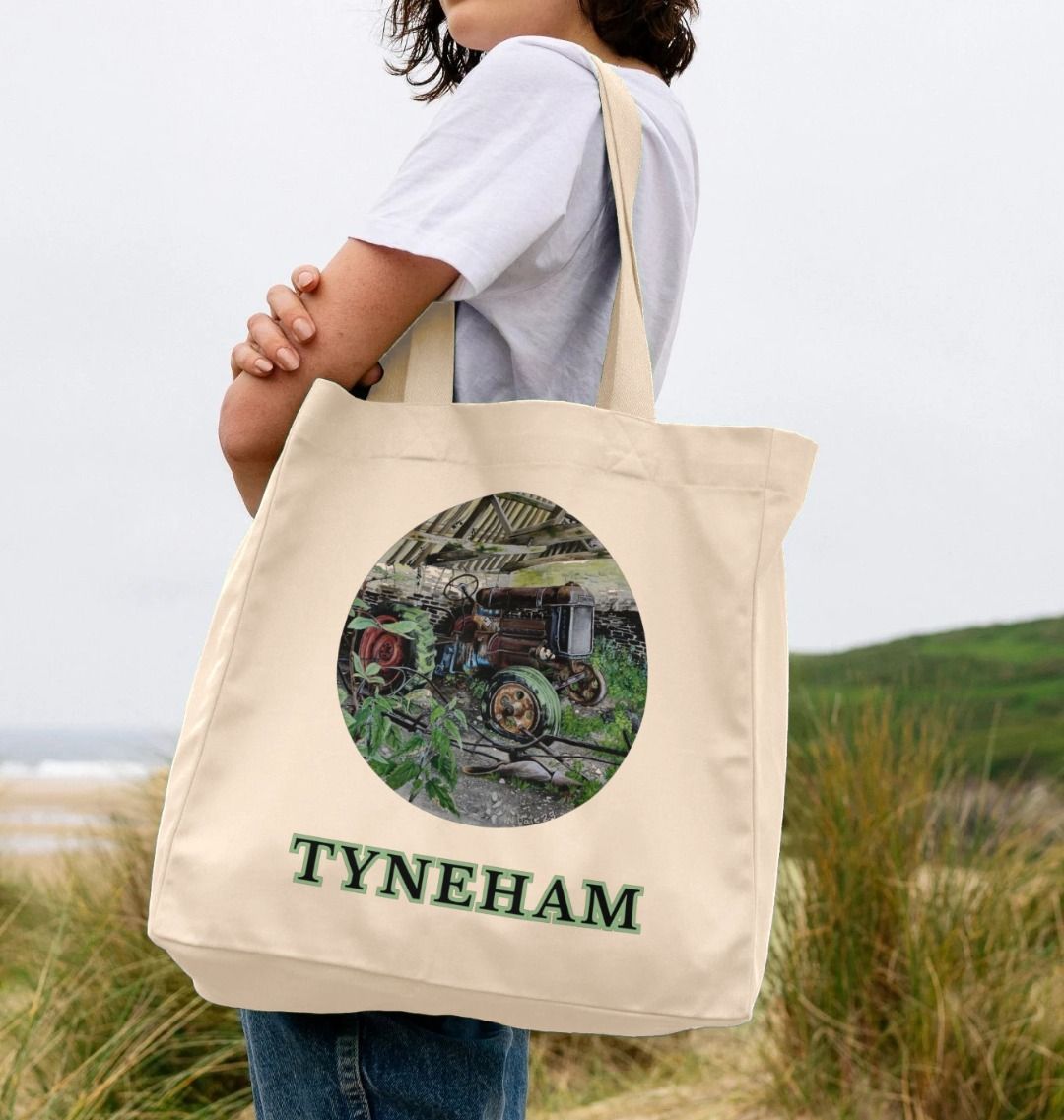 Image of a female model holding a Natural Shopper Tote Bag featuring a copy of the painting 'A Well Deserved Rest At Tyneham' by Nick Dale. The bag depicts a Fordson Major tractor laid up in a farm building at the abandoned village of Tyneham in Dorset.