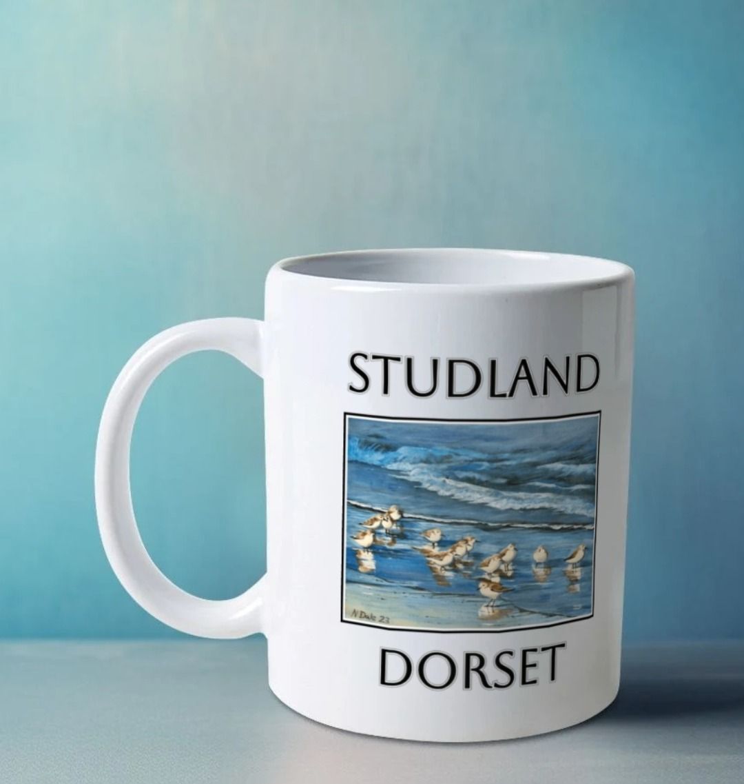 Image of a White Mug on a white table featuring a copy of the painting 'Studland Misty Morning' by Nick Dale, with the word 'Studland' written above the picture and 'Dorset' written beneath it. The handle is orientated to the left. The mug depicts Dunlins on Studland Beach (Dorset) on a crisp and misty morning.