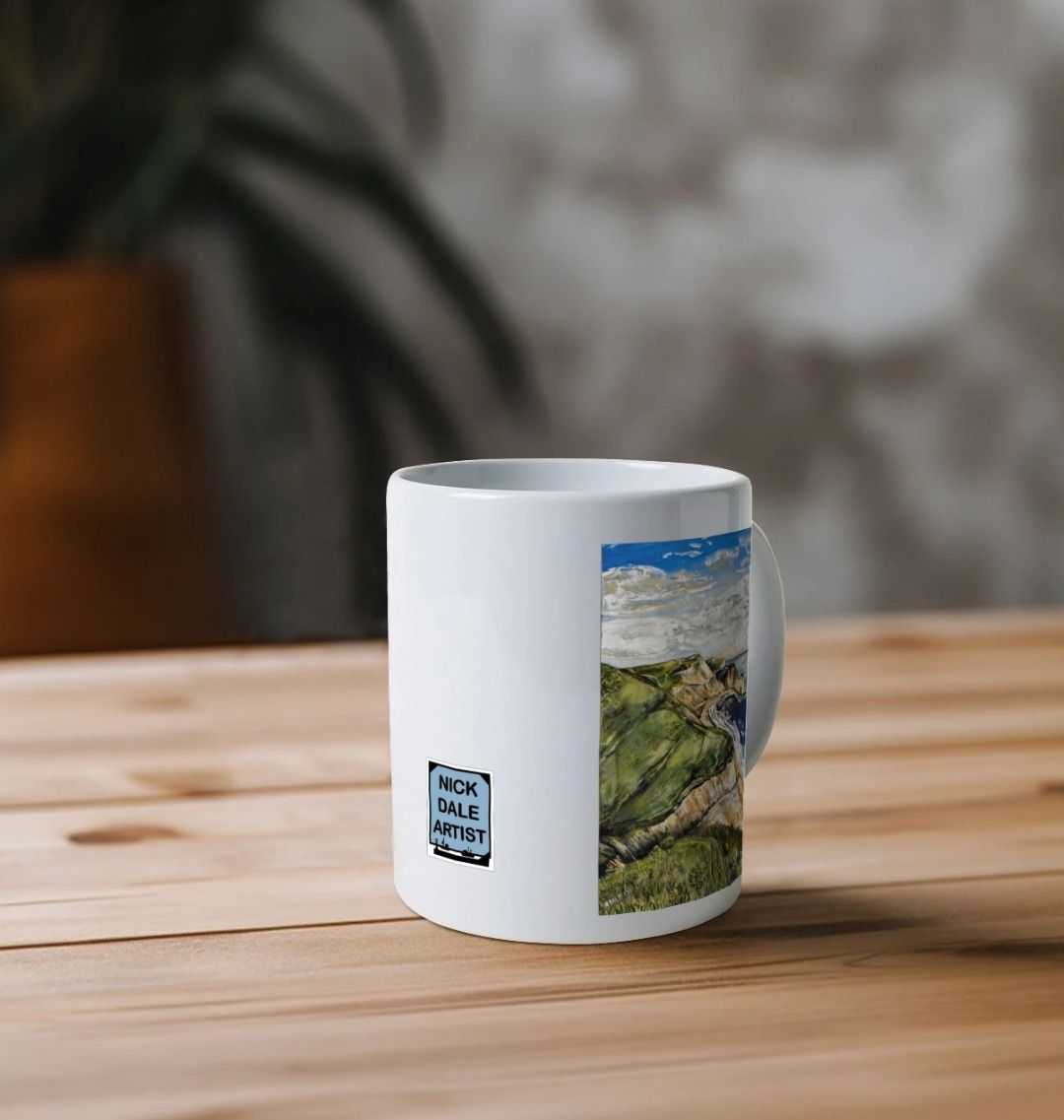 Image of a White Mug on a table featuring a copy of the painting 'Durdle Door From The West' by Nick Dale. Image also shows the artist's logo on the mug. The mug handle is obscured behind the mug. The mug depicts a view along the Jurassic Coast to the famous Dorset limestone arch landmark, as seen from the coastal path to the west.