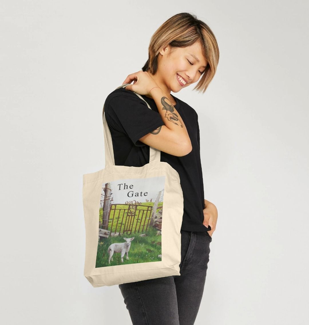 Image of a female model carrying a Natural Tote Bag featuring a copy of a detail from the painting 'The Gate' by Nick Dale, with the words 'the Gate' written above the gate. The bag depicts a lamb in front of an ornamental garden gate reused by a farmer somewhere in North Devon, probably to the south of Valley of Rocks, Lynton. The lamb was observed on Lundy Island, Devon.