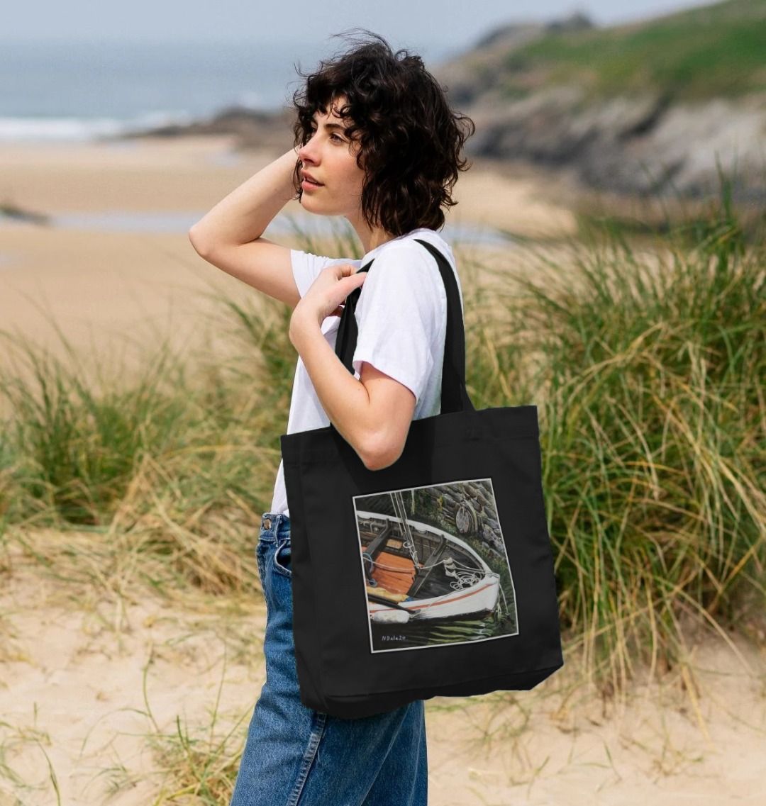 Image of a female model carrying a Black Shopper Tote Bag featuring a copy of the painting 'The Outlet' by Nick Dale. The bag depicts a view down into the harbour at Lynmouth in North Devon showing a small sailing boat at rest against the harbour wall next to a water outfall pipe.