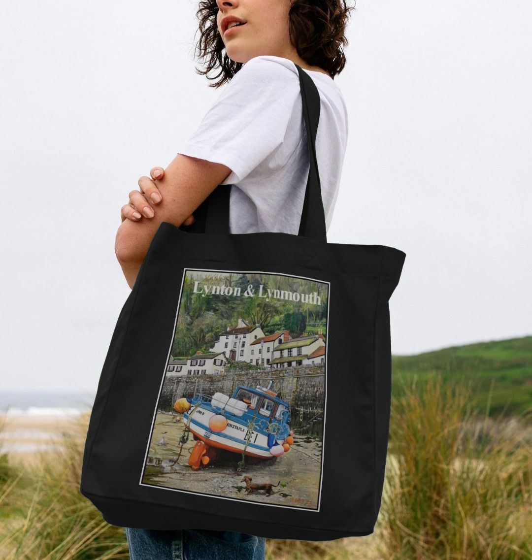 Image of a female model carrying a Black Shopper Tote Bag featuring a copy of the painting 'Lynmouth At Lowest Tide' by Nick Dale, with the words 'Lynton & Lynmouth' written across the top. The bag depicts an unusual perspective on the picturesque harbour of Lynmouth (Devon), as seen from the floor of the harbour at low tide. Behind the fishing boat can be seen the characterful harbourside buildings.