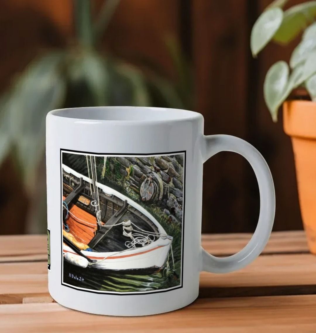 Image of a White Mug on a wooden shelf featuring a copy of the painting 'The Outlet' by Nick Dale. The handle is orientated to the right. The mug depicts a view down into the harbour at Lynmouth in North Devon showing a small sailing boat at rest against the harbour wall next to a water outfall pipe.