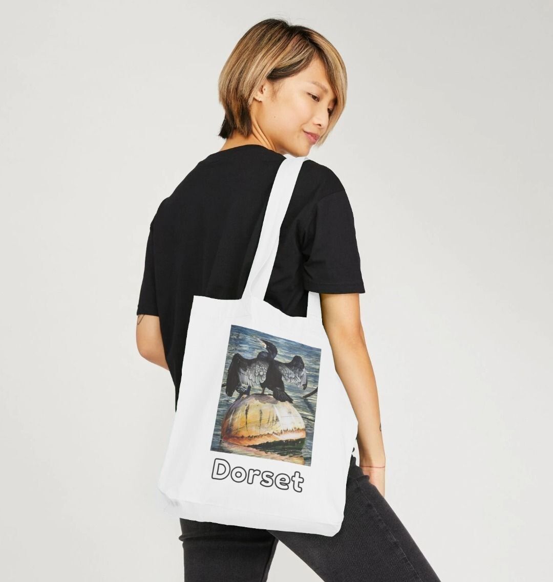 Image of a female model holding a White Tote Bag featuring a modified copy of the painting 'Cormorant At West Bay' by Nick Dale. The bag depicts a close-up of a cormorant sunbathing on a mooring buoy in West Bay's harbour in Dorset.