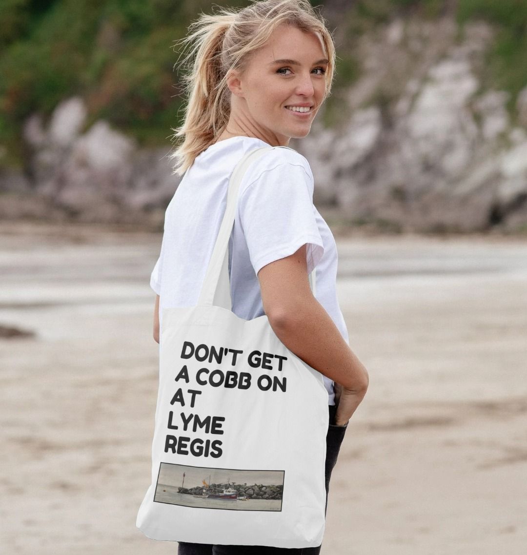 Image of a female model carrying a White Tote Bag featuring a copy of the painting 'End Of The Line' by Nick Dale with the slogan 'Don't Get A Cobb On At Lyme Regis' written above it. The bag depicts a fisherman preparing his catch for unloading at Lyme Regis as the birds line up on the breakwater and patiently await their turn to feed.