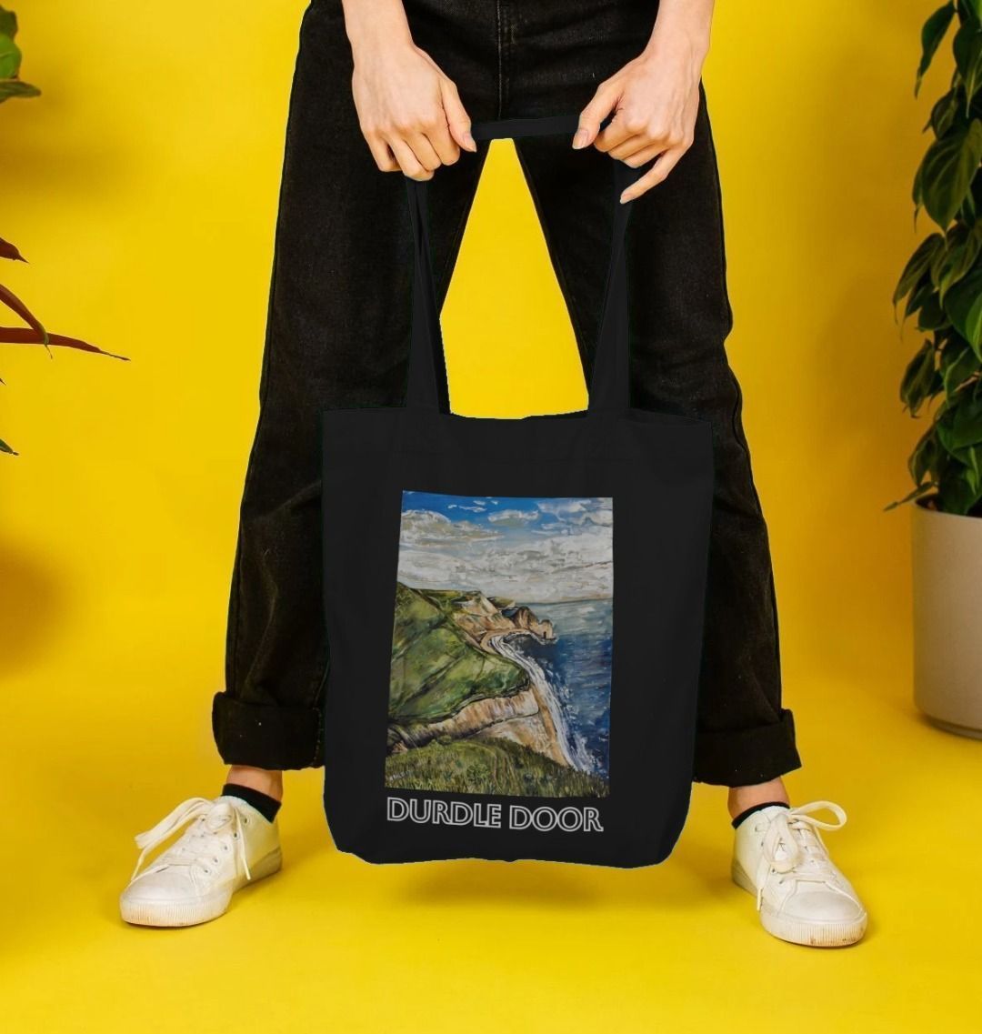 Image of female model carrying a Black Tote Bag featuring a copy of the painting 'Durdle Door From The West' by Nick Dale with the words 'Durdle Door' written below it. The bag depicts a view along the Jurassic Coast to the famous Dorset limestone arch landmark, as seen from the coastal path to the west.