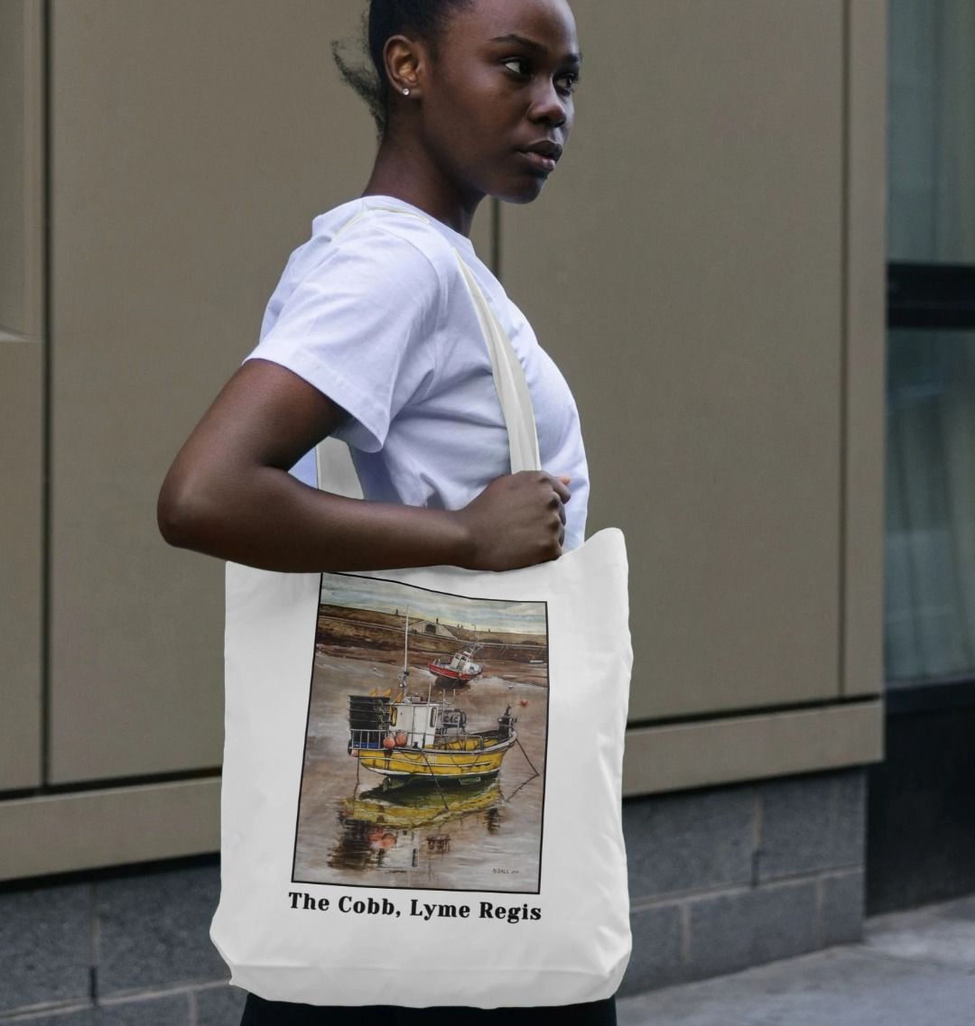 Image of a female model carrying a White Tote Bag featuring a copy of the painting 'Lyme Regis at Low Tide' by Nick Dale, with the words 'The Cobb, Lyme Regis' written beneath it. The bag depicts a sunset where the fishing vessels rest on the floor of Lyme Regis harbour whilst the tourists enjoy the view from the Cobb.
