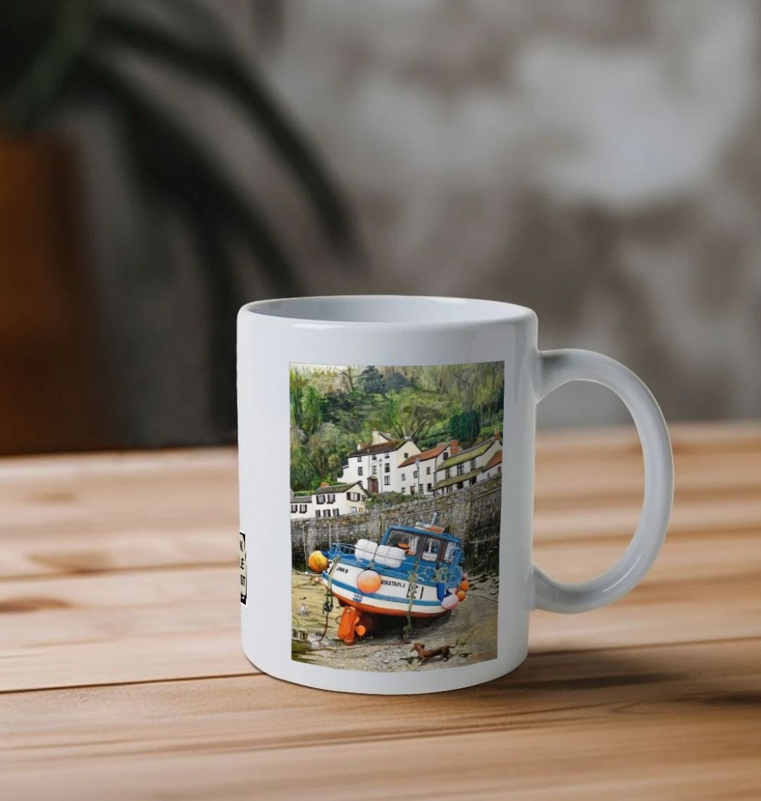 Image of a White Mug on a wooden table featuring a copy of the painting 'Lynmouth At Lowest Tide' by Nick Dale. The handle is orientated to the right. The mug depicts an unusual perspective on the picturesque harbour of Lynmouth (Devon), as seen from the floor of the harbour at low tide. Behind the fishing boat can be seen the characterful harbourside buildings.