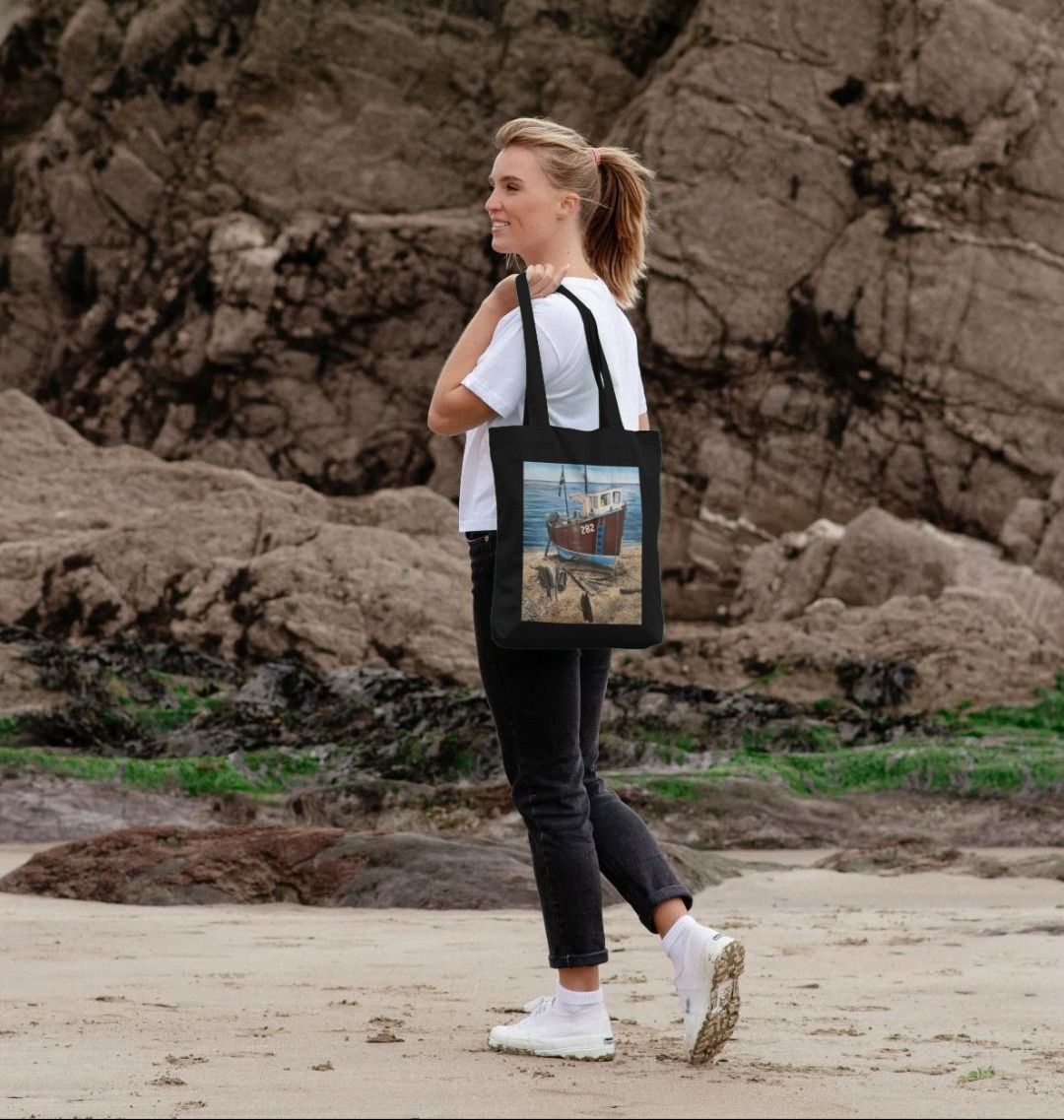 Image of female model carrying a Black Tote Bag featuring a copy of the painting 'Relaxing Beer' by Nick Dale. The bag depicts a fishing boat beached at Beer, Devon. The wood is laid under the boats to aid in their winching up the shingle beach.
