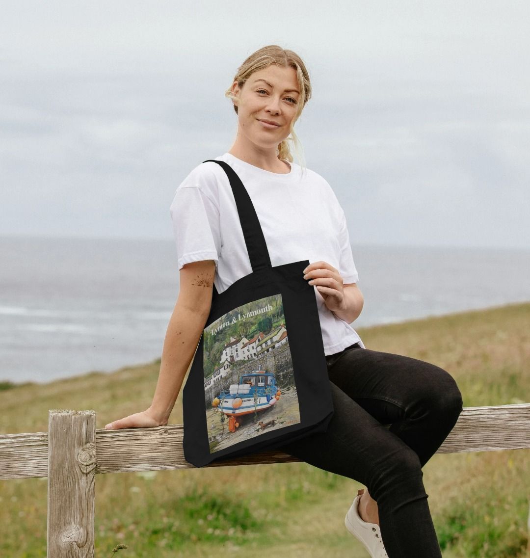 Image of a female model carrying a Black Tote Bag featuring a copy of the painting 'Lynmouth At Lowest Tide' by Nick Dale, with the words 'Lynton & Lynmouth' written across the top. The bag depicts an unusual perspective on the picturesque harbour of Lynmouth (Devon), as seen from the floor of the harbour at low tide. Behind the fishing boat can be seen the characterful harbourside buildings.