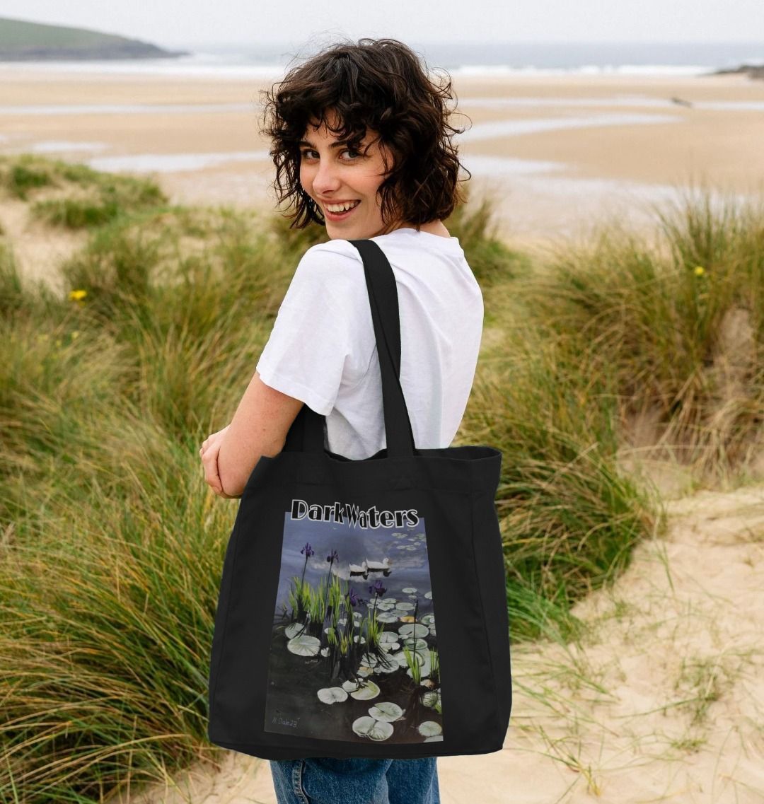 Image of female model carrying a Black Shopper Tote Bag featuring a copy of the painting 'Dark Waters' by Nick Dale, with the words 'Dark Waters' written above it. The bag depicts lily pads, irises and ducks on a pond at Knightshayes Court, Devon.