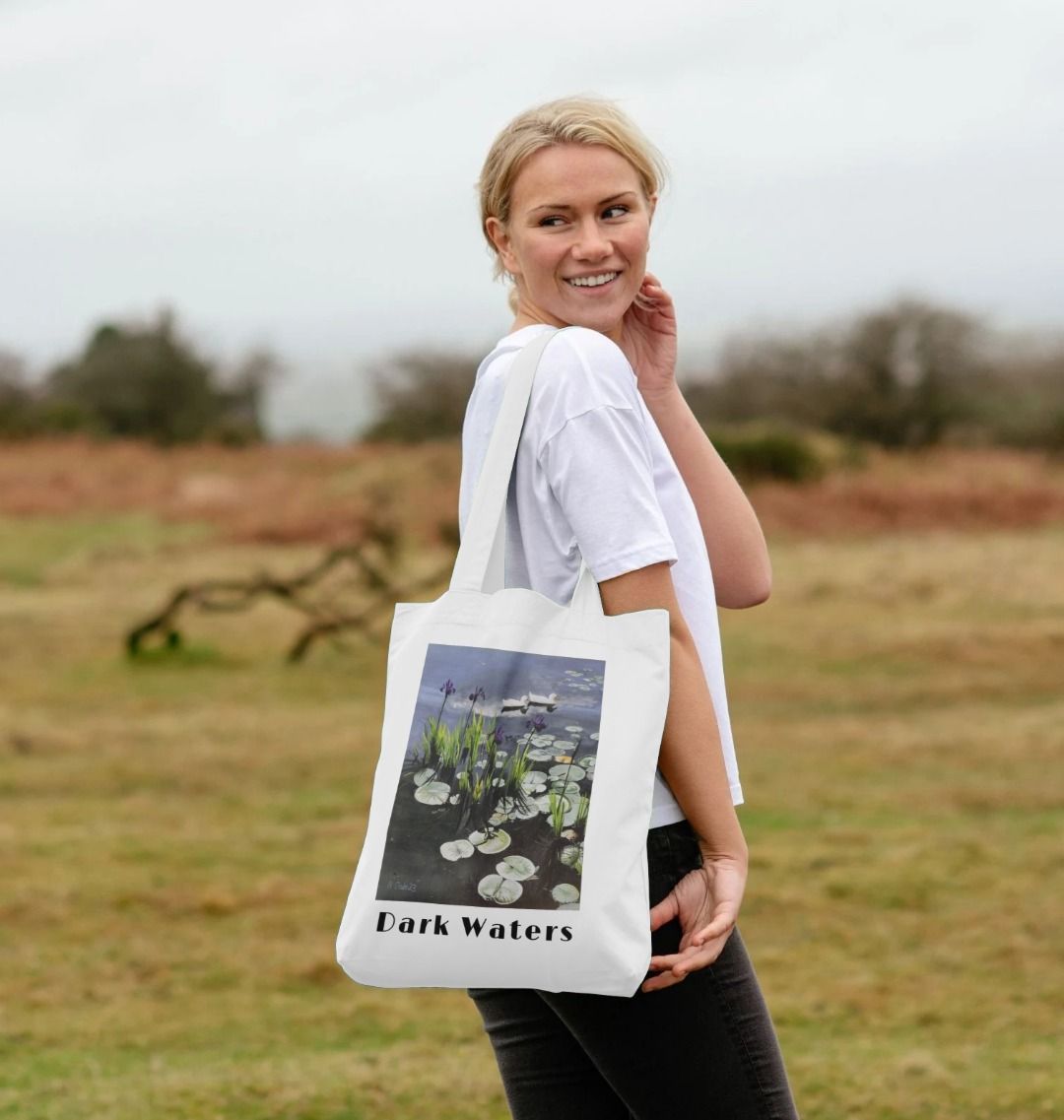 Image of a female model carrying a White Tote Bag featuring a copy of the painting 'Dark Waters' by Nick Dale, with the words 'Dark Waters' written beneath it. The bag depicts lily pads, irises and ducks on a pond at Knightshayes Court, Devon.