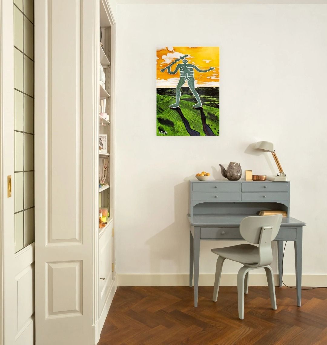 Image of a Large Art Print featuring a copy of the painting 'Cerne Abbas Giant Pootling Along' by Nick Dale; as displayed on a wall above a desk. The print imagines the Cerne Abbas Giant chalk hill figure wandering across the Dorset landscape. The giant can be seen scaling Hod Hill in the Blackmore Vale.