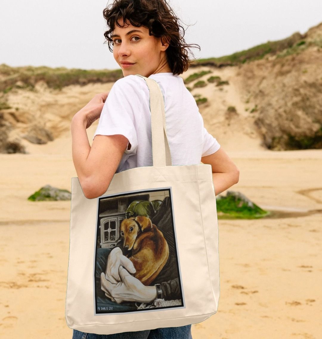 Image of a female model holding a Natural Shopper Tote Bag featuring a copy of the painting 'Amber Concordia' by Nick Dale. The bag depicts a dog sitting curled-up on the lap of its owner in front of the hearth.