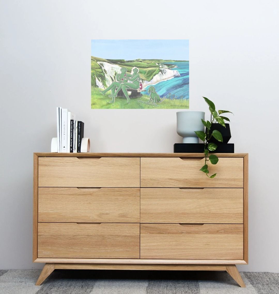 Image of a Large Art Print (A3 size) featuring a copy of the composite painting 'Mr & Mrs Cerne Abbas Giant's Picnic At White Nothe' by Nick Dale; as displayed on a wall above a chest of drawers. The print depicts Mr & Mrs Cerne Abbas Giant having a merlot picnic whilst on a date along the Dorset coast at the White Nothe, east of Weymouth. They are joined on the date by their cocker spaniel dog.