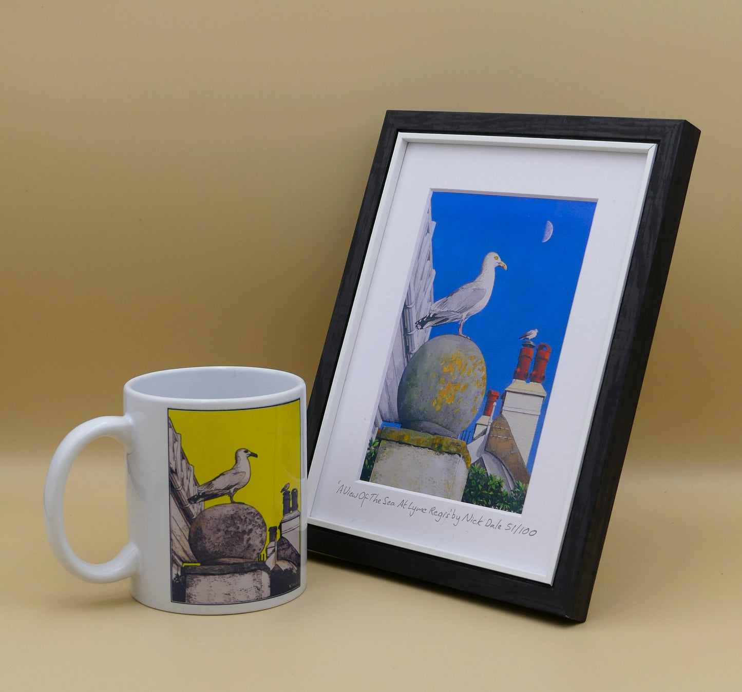 Image of Small Art Print featuring a copy of the painting 'A View Of The Sea At Lyme Regis' by Nick Dale. The print depicts a gull and buildings along Marine Parade, Lyme Regis. The print is situated in a black frame next to a mug.