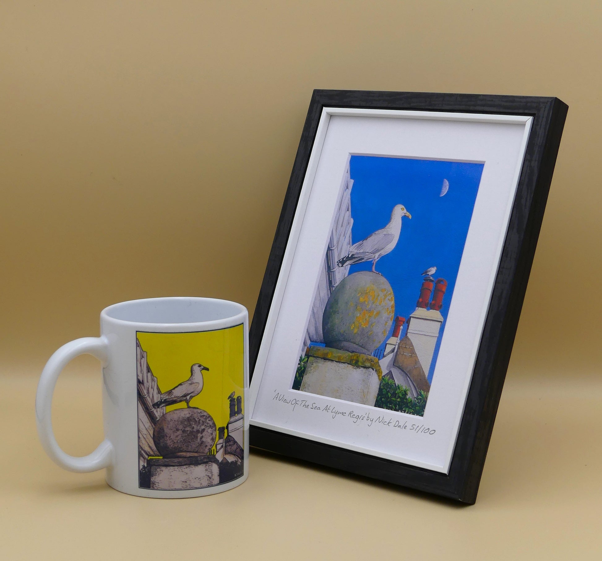 Image of Small Art Print featuring a copy of the painting 'A View Of The Sea At Lyme Regis' by Nick Dale. The print depicts a gull and buildings along Marine Parade, Lyme Regis. The print is situated in a black frame next to a mug.