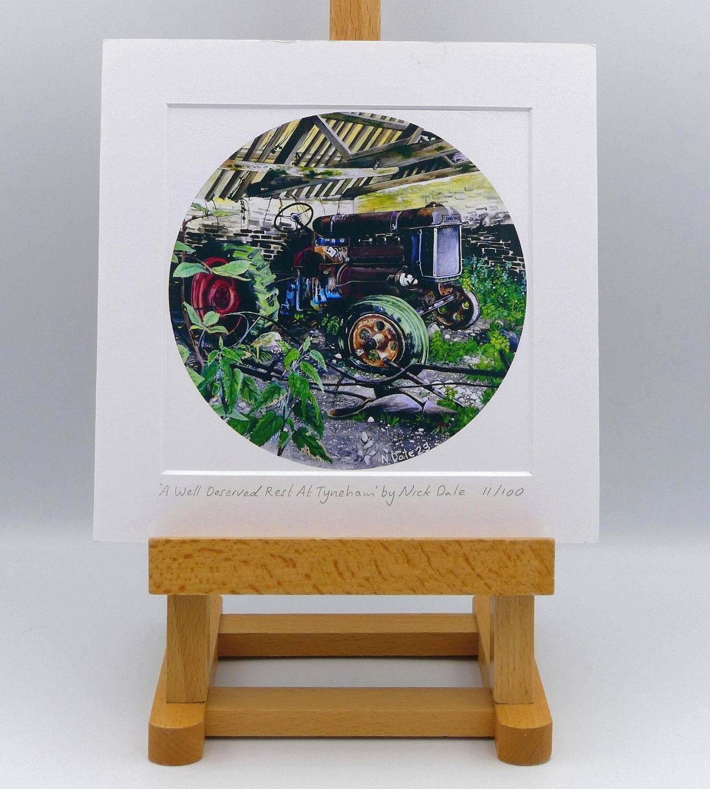 Image of Square Medium Art Print featuring a copy of the painting 'A Well Deserved Rest At Tyneham' by Nick Dale. The print depicts a Fordson Major tractor laid up in a farm building at the abandoned village of Tyneham in Dorset. The print is situated on a small easel.