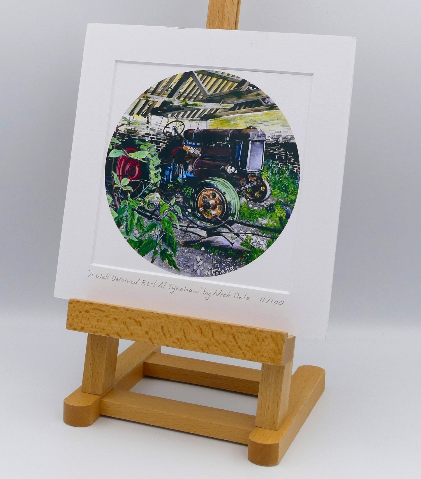 Image of Square Medium Art Print featuring a copy of the painting 'A Well Deserved Rest At Tyneham' by Nick Dale. The print depicts a Fordson Major tractor laid up in a farm building at the abandoned village of Tyneham in Dorset. The print is situated on a small easel.