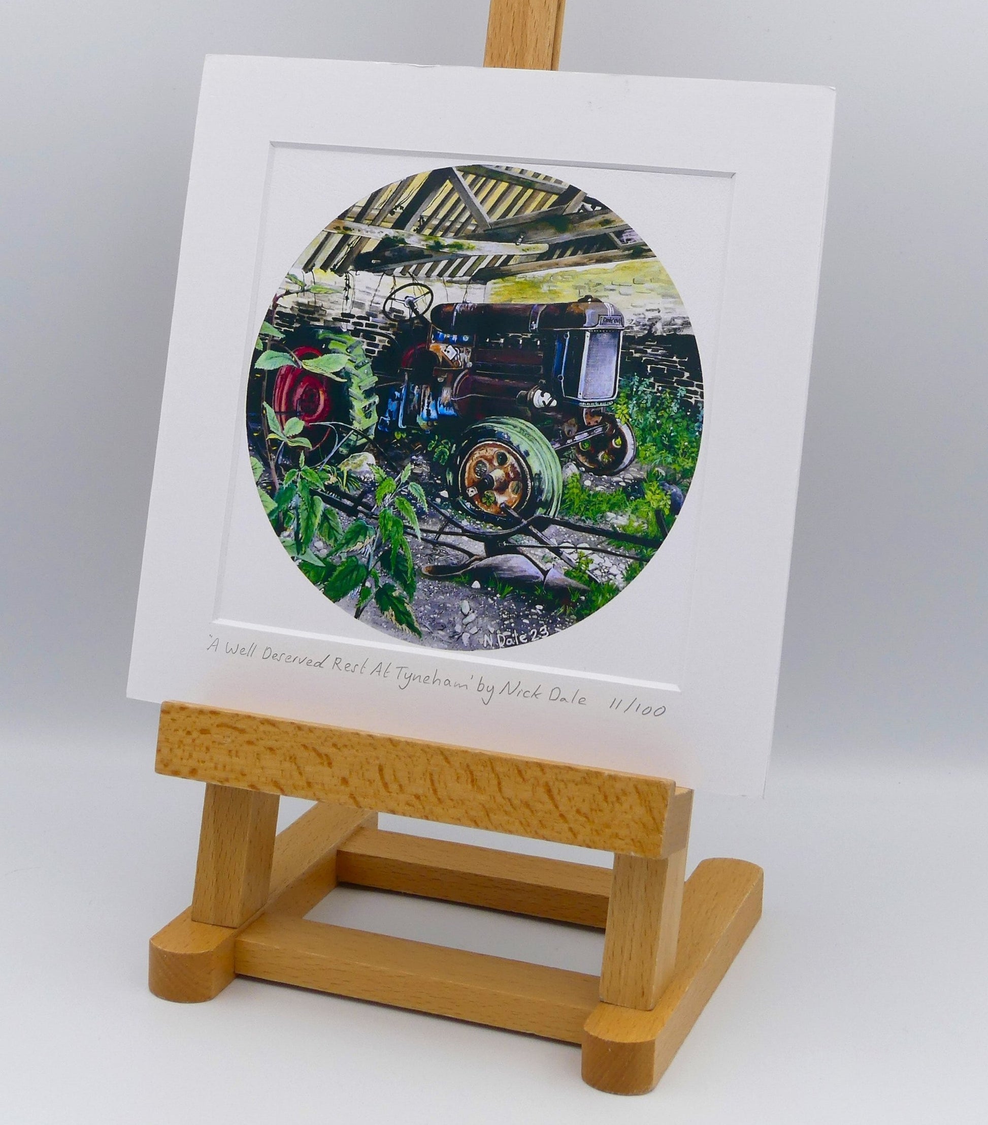 Image of Square Medium Art Print featuring a copy of the painting 'A Well Deserved Rest At Tyneham' by Nick Dale. The print depicts a Fordson Major tractor laid up in a farm building at the abandoned village of Tyneham in Dorset. The print is situated on a small easel.