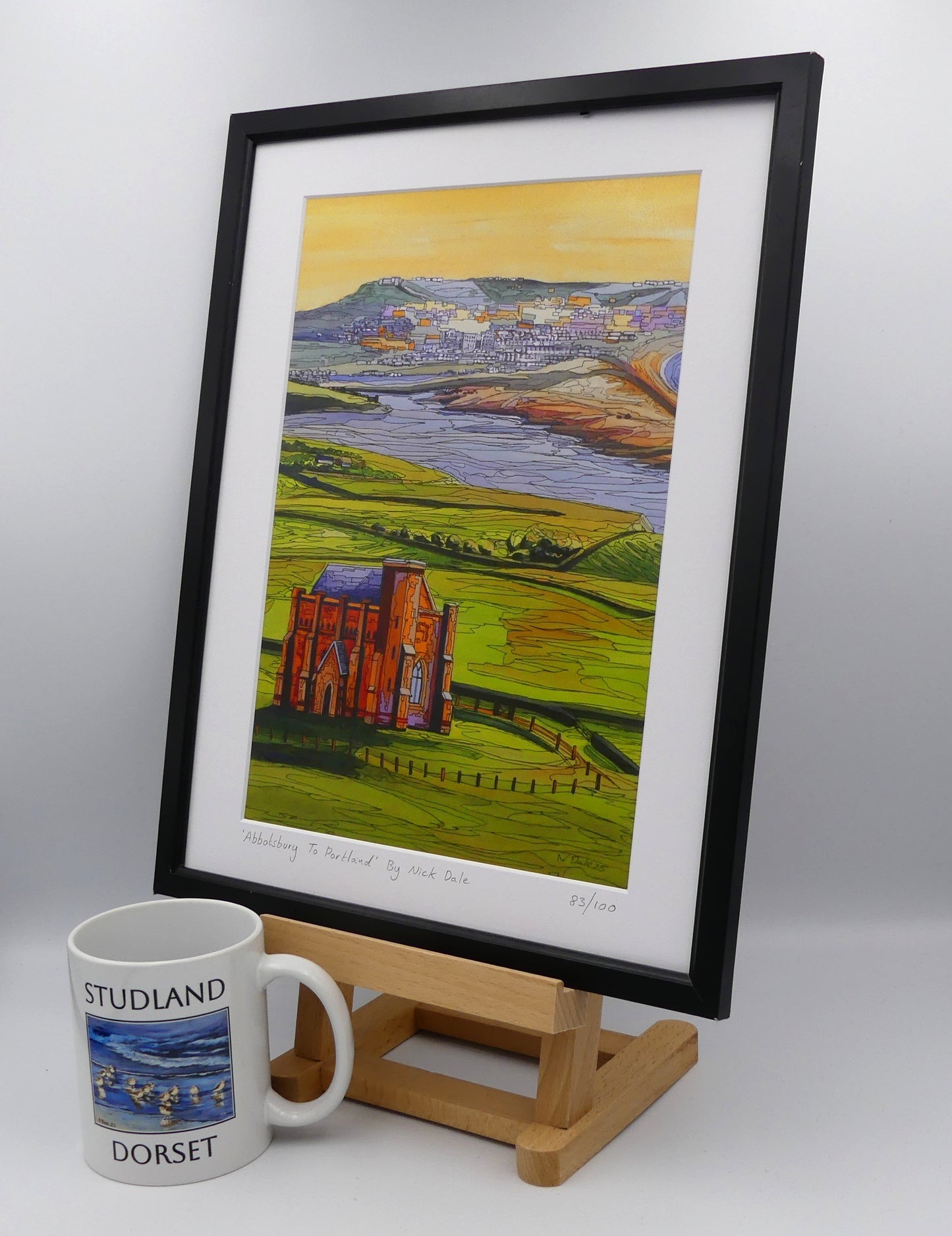 Image of a Medium Art Print featuring a copy of the painting 'Abbotsbury To Portland' by Nick Dale. The print depicts the view along the Dorset coast from St Catherine's Chapel above Abbotsbury, across The Fleet lagoon to Chesil Beach and ultimately to Portland beyond it. The print is situated on a small easel in a black frame next to a mug.