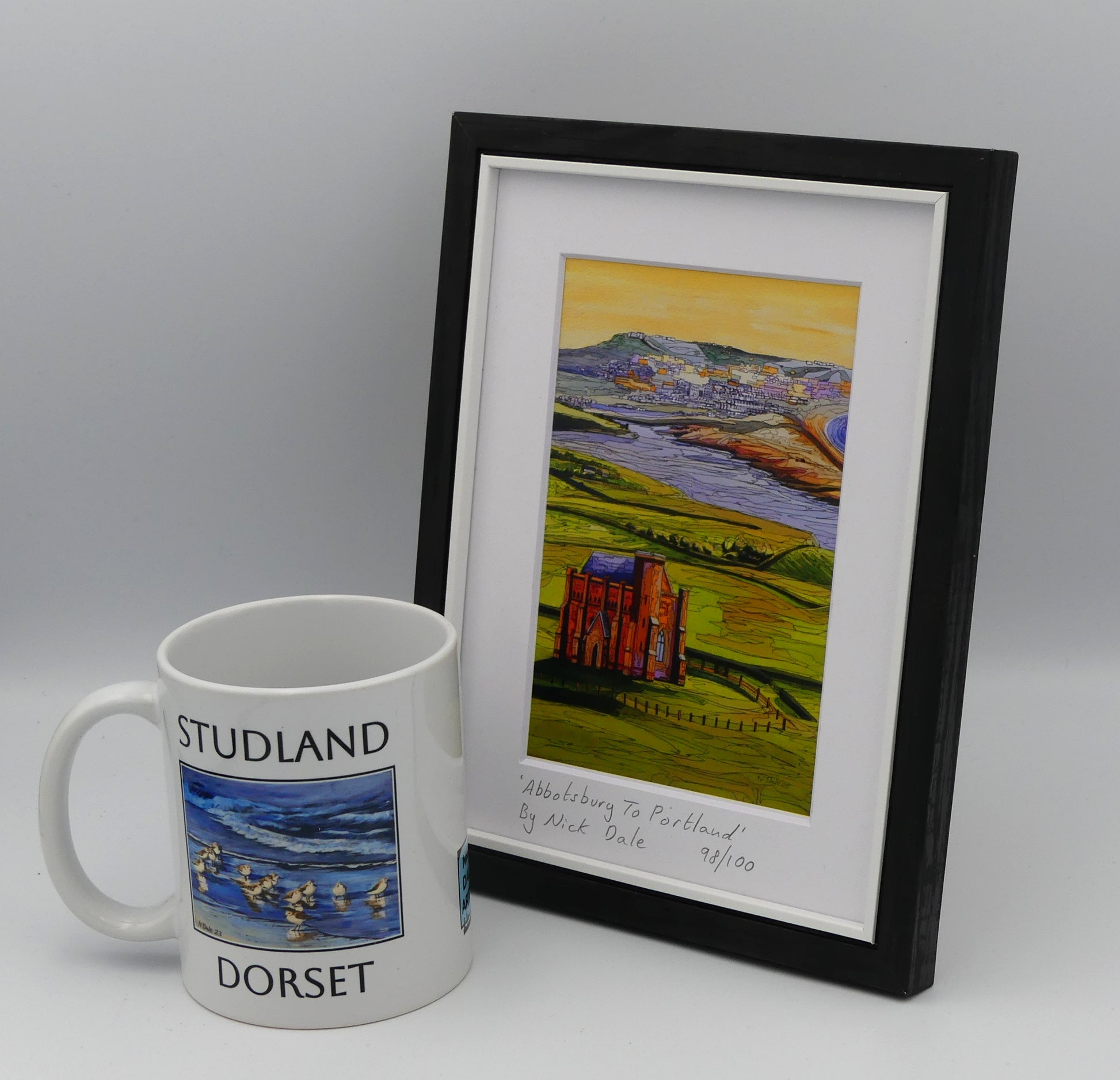 Image of a Small Art Print featuring a copy of the painting 'Abbotsbury To Portland' by Nick Dale. The print depicts the view along the Dorset coast from St Catherine's Chapel above Abbotsbury, across The Fleet lagoon to Chesil Beach and ultimately to Portland beyond it. The print is situated in a black frame next to a mug.