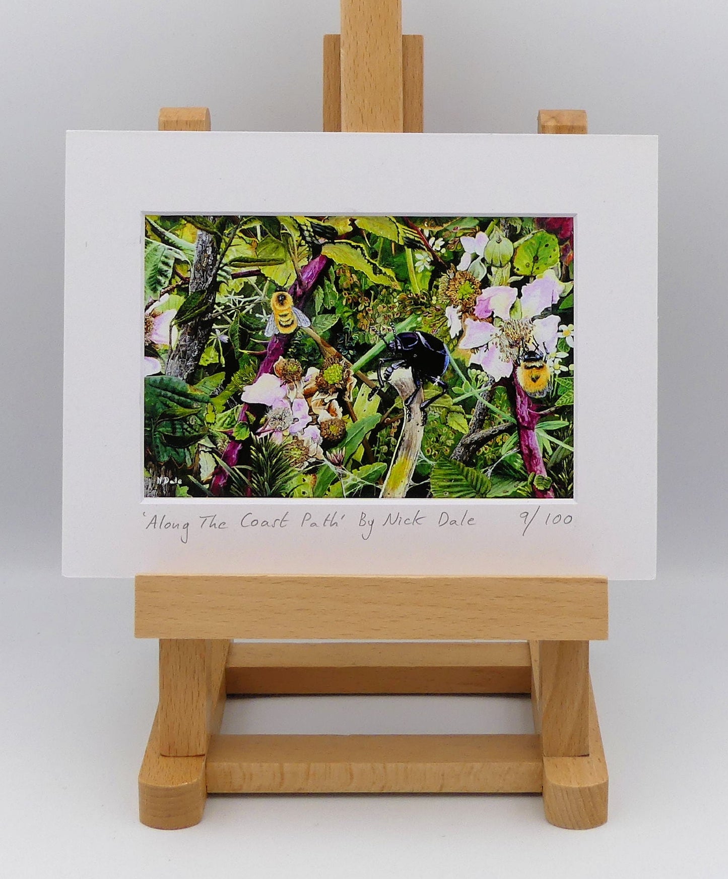 Image of a Small Art Print featuring a copy of the painting 'Along The Coast Path' by Nick Dale. The print depicts a close-up view of a coast path hedgerow on the Dorset/Devon border. A bloody-nosed beetle edges its way along a plant stem whilst bees attend the bramble flowers. The print is situated on a small easel.