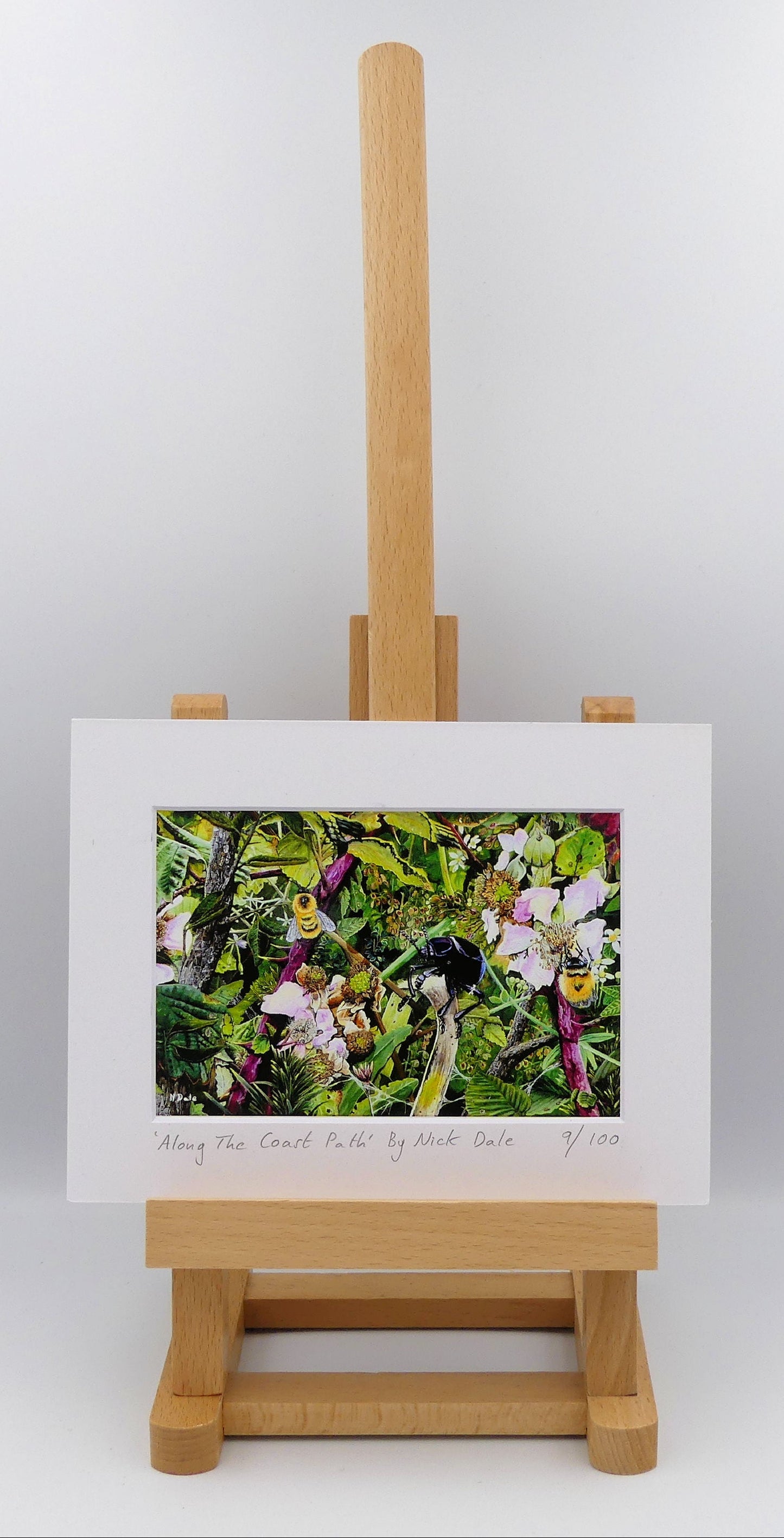 Image of a Small Art Print featuring a copy of the painting 'Along The Coast Path' by Nick Dale. The print depicts a close-up view of a coast path hedgerow on the Dorset/Devon border. A bloody-nosed beetle edges its way along a plant stem whilst bees attend the bramble flowers. The print is situated on a small easel.