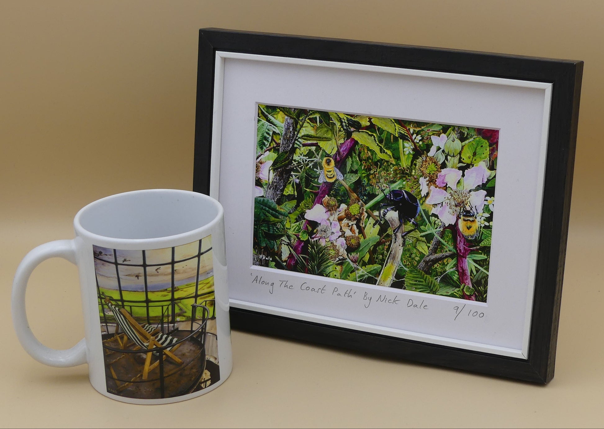 Image of a Small Art Print featuring a copy of the painting 'Along The Coast Path' by Nick Dale. The print depicts a close-up view of a coast path hedgerow on the Dorset/Devon border. A bloody-nosed beetle edges its way along a plant stem whilst bees attend the bramble flowers. The print is situated in a black frame next to a mug.
