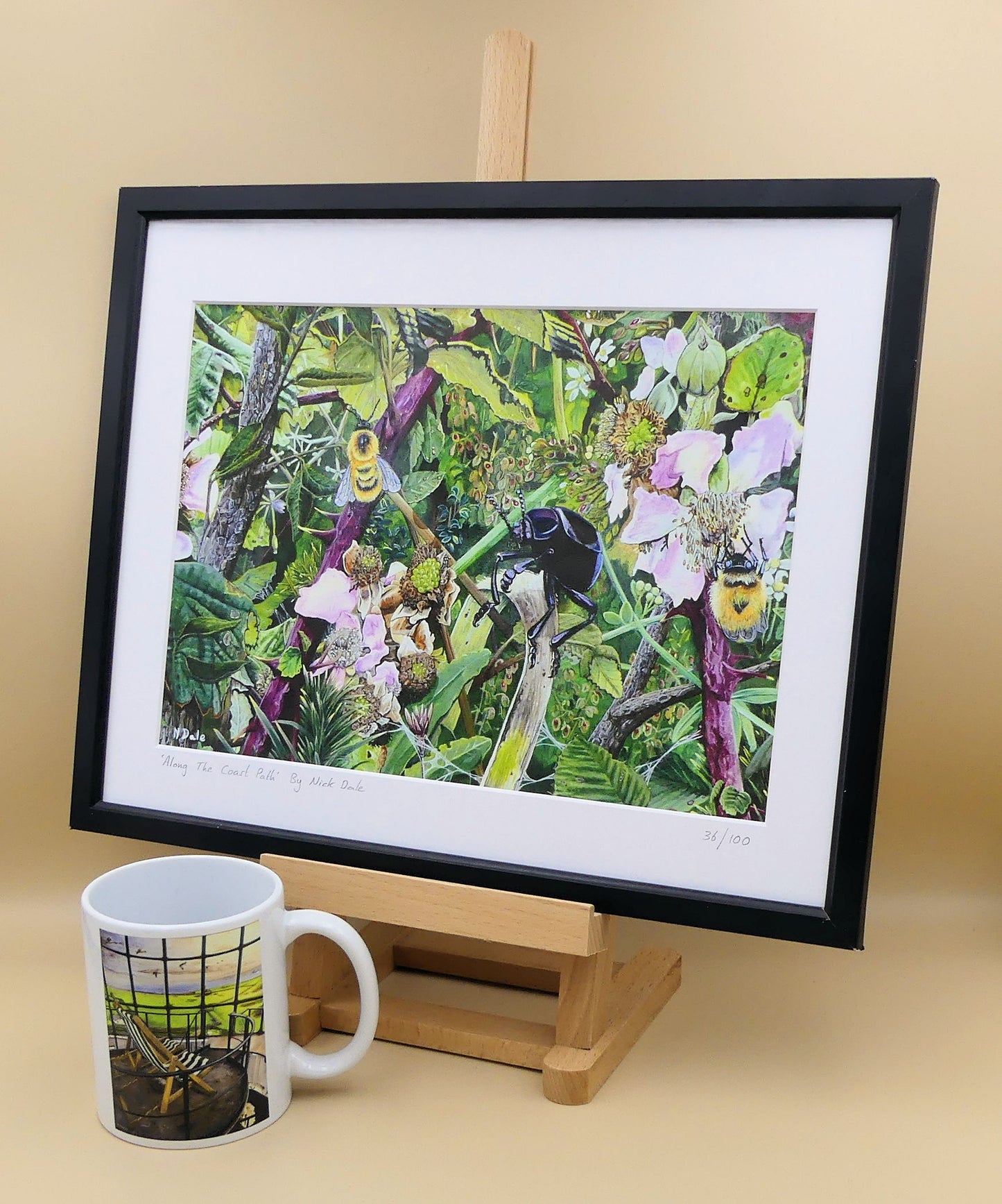 Image of a Medium Art Print featuring a copy of the painting 'Along The Coast Path' by Nick Dale. The print depicts a close-up view of a coast path hedgerow on the Dorset/Devon border. A bloody-nosed beetle edges its way along a plant stem whilst bees attend the bramble flowers. The print is situated on a small easel in a black frame next to a mug.