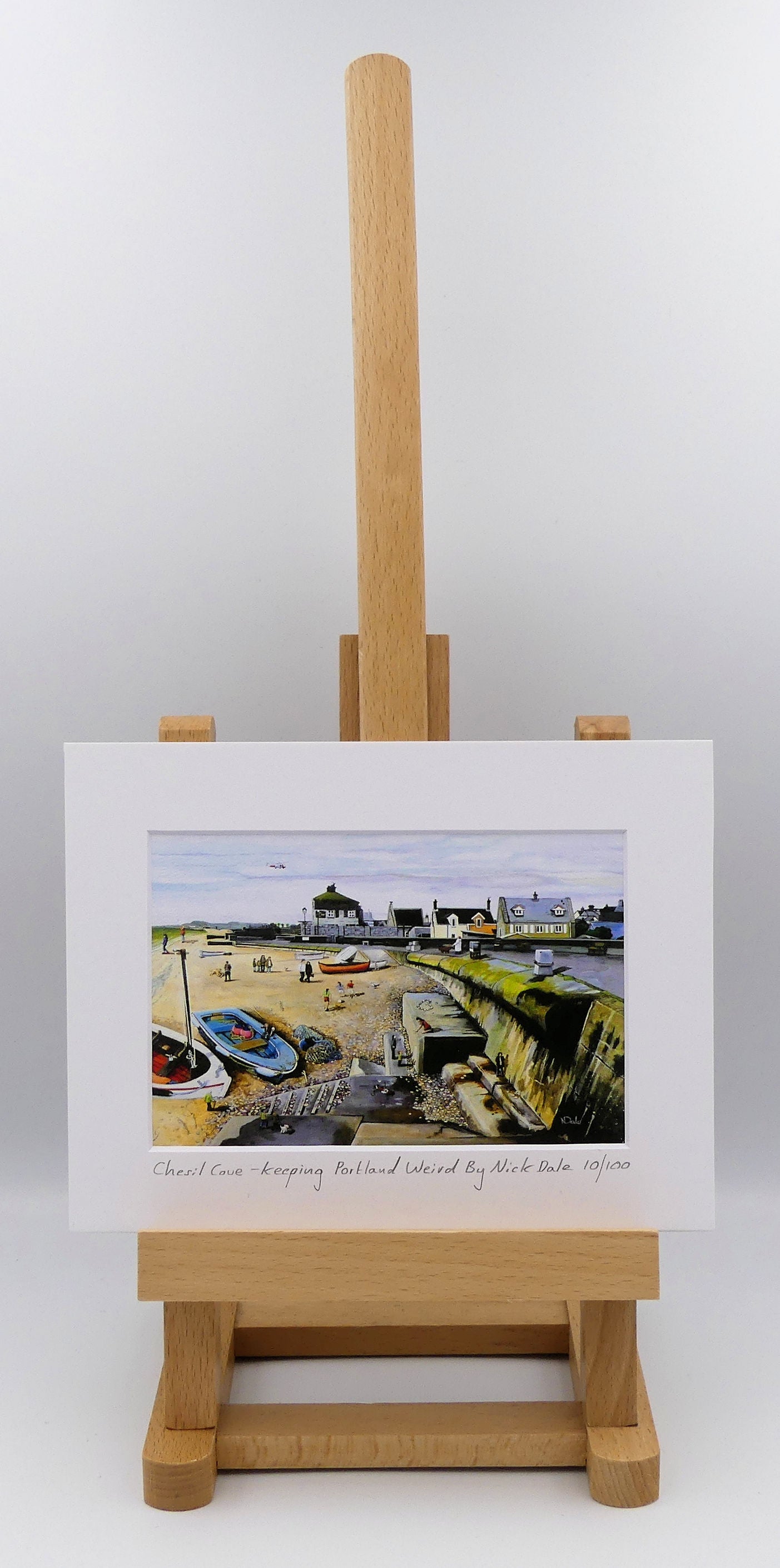 Image of a Small Art Print featuring a copy of the painting 'Chesil Cove - Keeping Portland Weird' by Nick Dale. The print depicts the end of Chesil Beach where it meets Portland at Chesil Cove. In the scene the size of the people is not affected by their distance from the foreground, hence making their appearance slightly surreal. The print is situated on a small easel.
